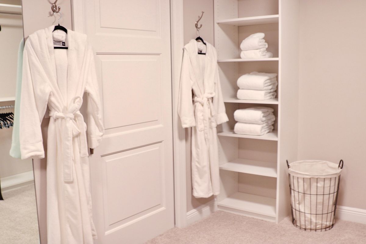 White bathrobes hang next to a closet with towels, and a laundry basket sits nearby.
