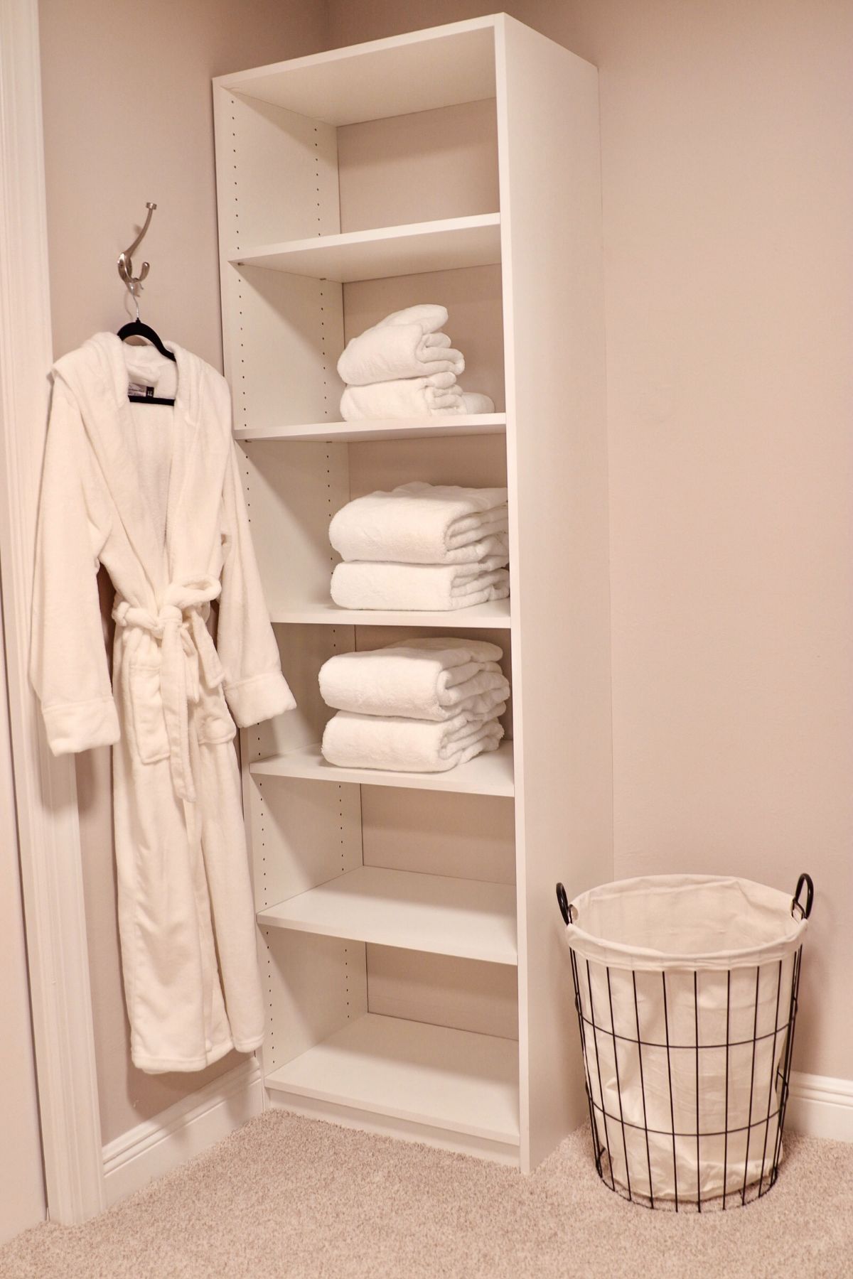 White shelving unit with folded towels, a robe on a hook, and a laundry basket.