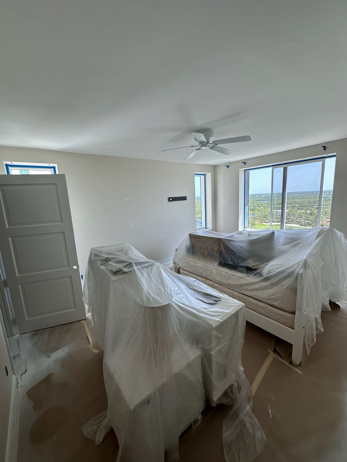 A room with furniture covered in plastic sheeting. White walls, doors, and a ceiling fan.