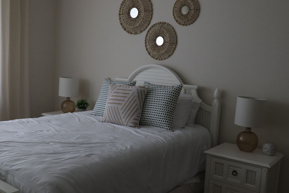 Bedroom with white bed, wicker headboard, bedside tables, lamps, and wall decor.