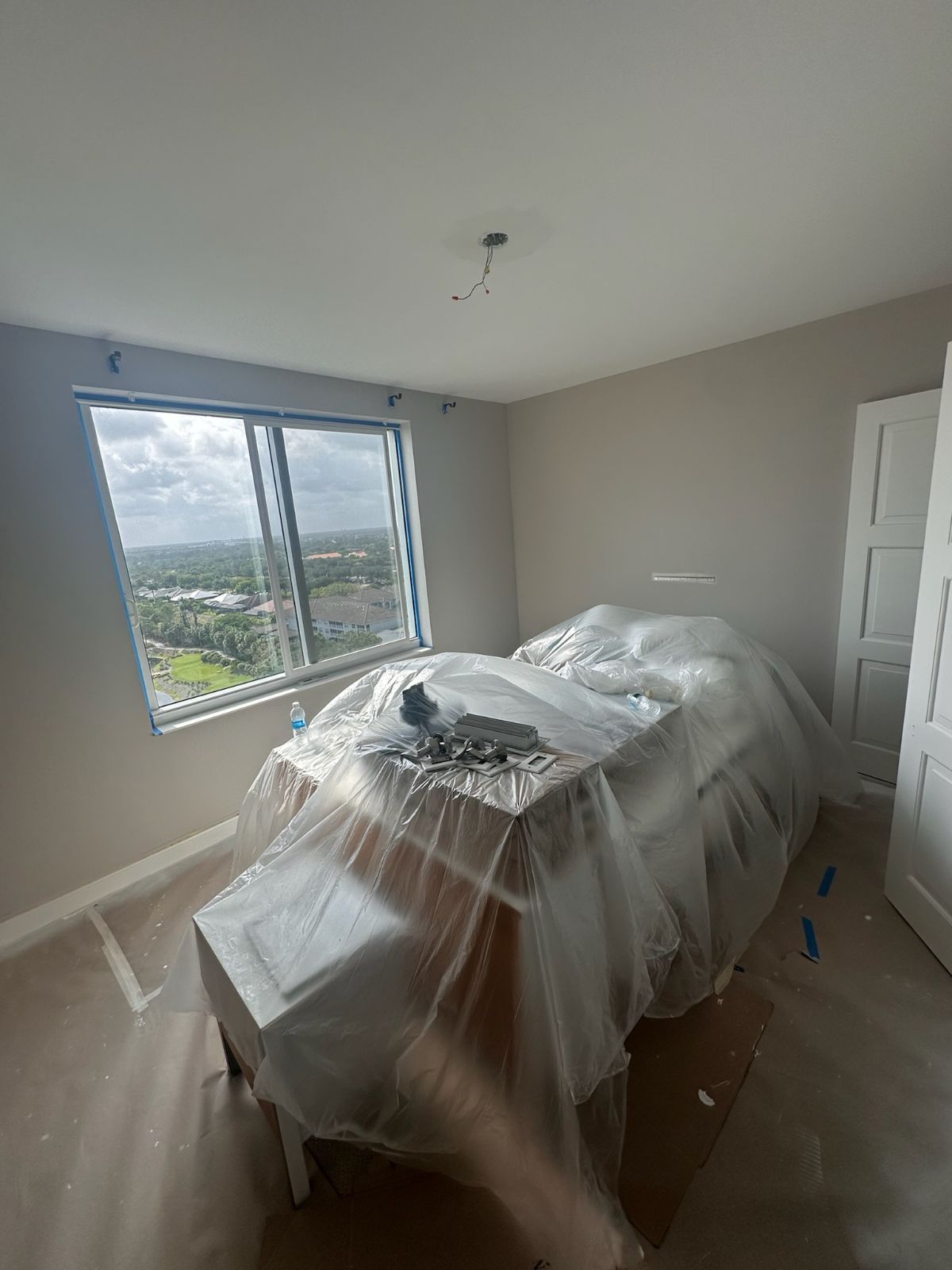A room with a covered table near a window, unfinished with neutral-toned walls and a light fixture.