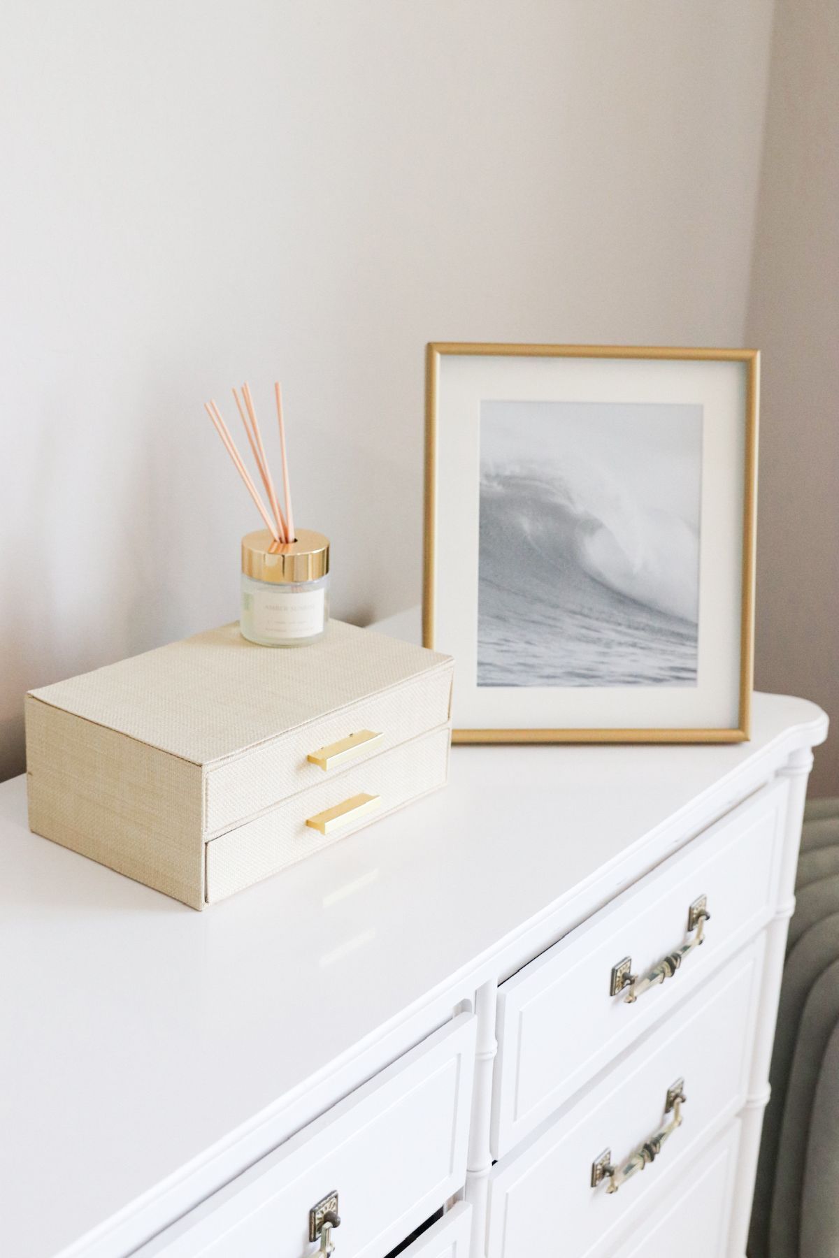 White dresser with gold accents, topped with a jewelry box, reed diffuser, and a framed ocean wave print.