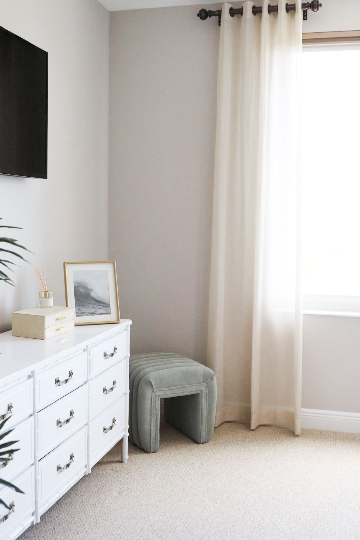 White dresser, framed art, and green ottoman in a bedroom corner, near a window with cream curtains.