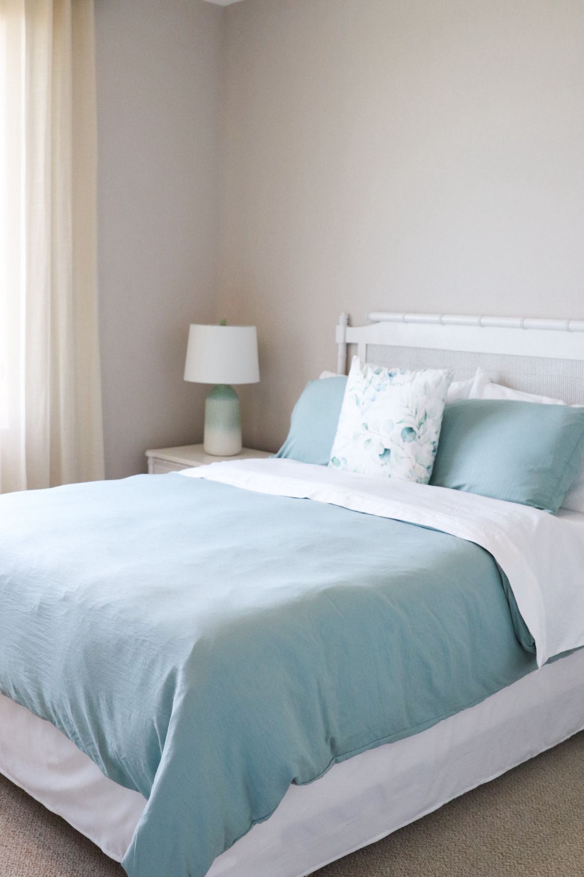 Bedroom with a white bed frame, light blue bedding, and a white nightstand with a teal lamp.