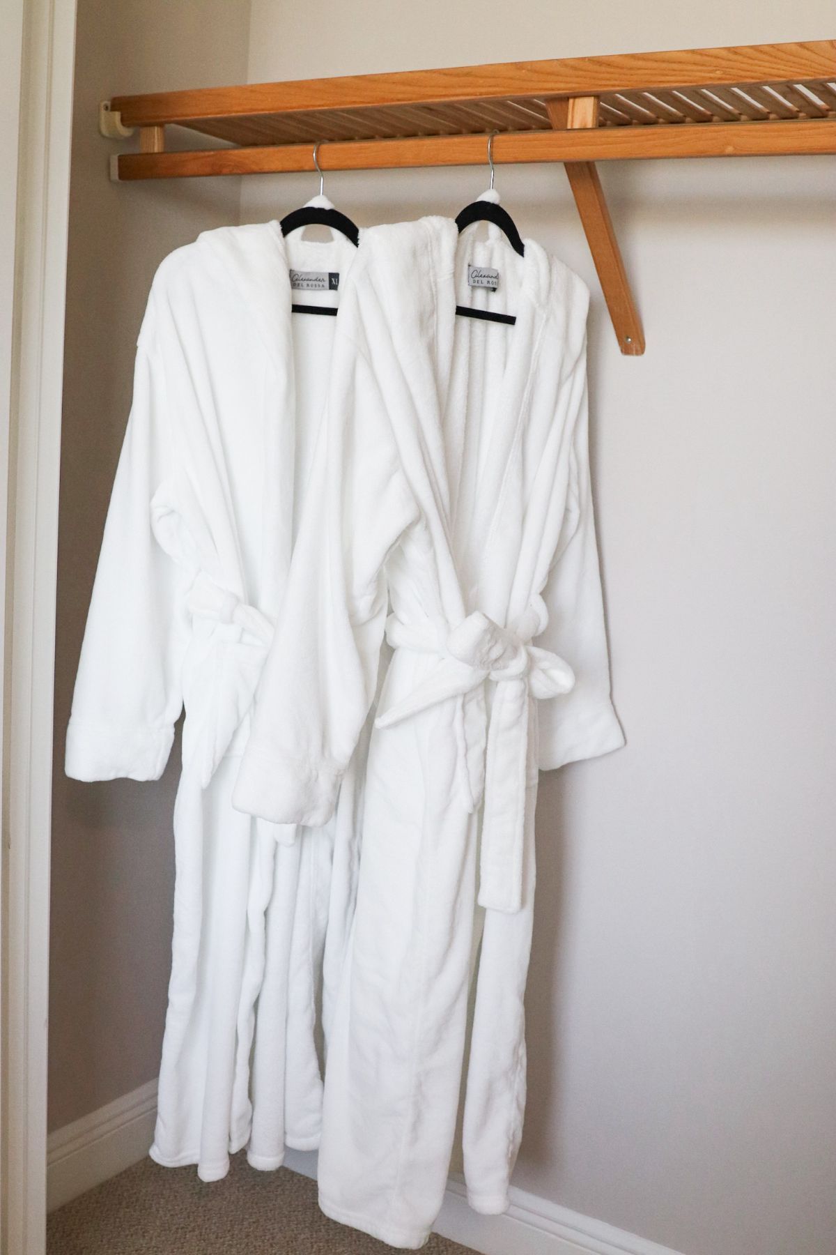 Two white bathrobes hanging on a wooden rack inside a closet.