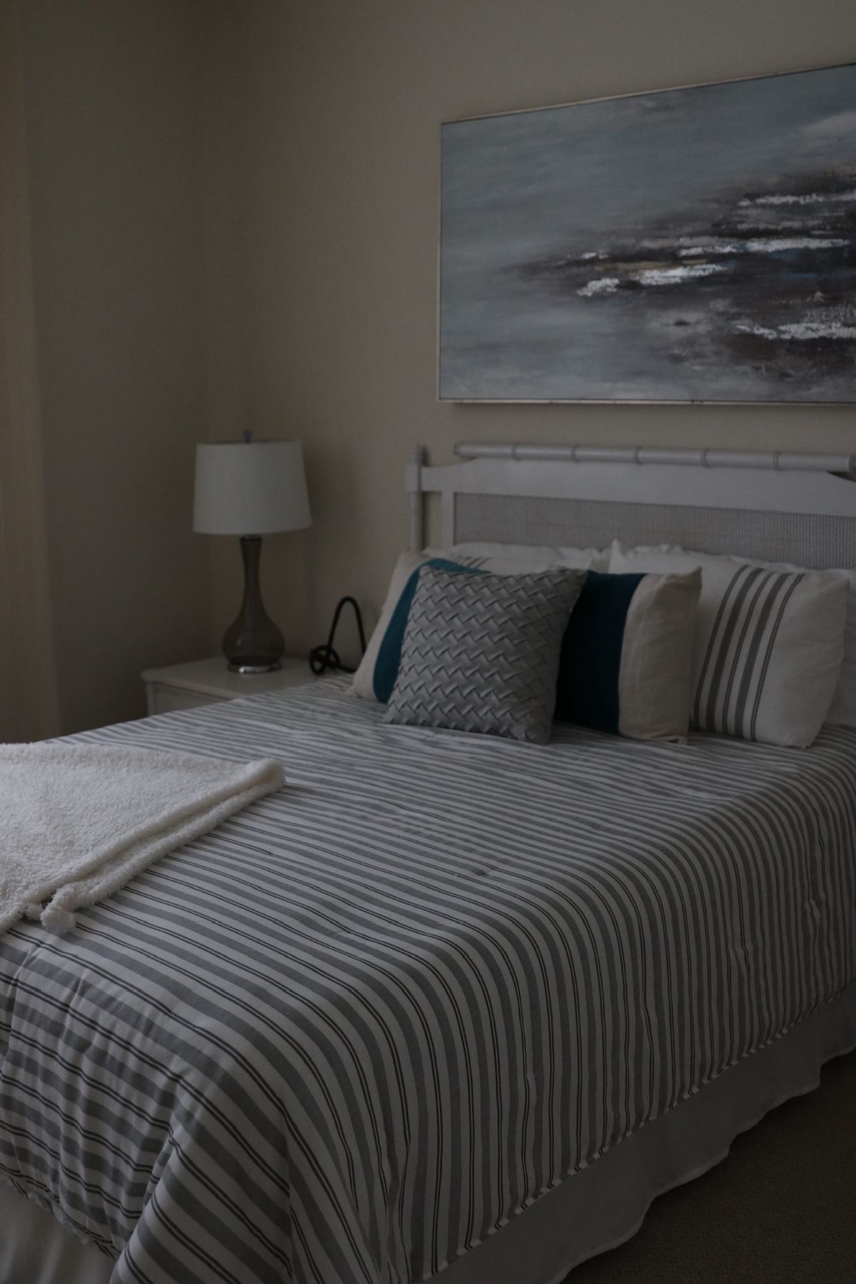 Bedroom with a striped quilt, pillows, lamp, and abstract art above the bed.