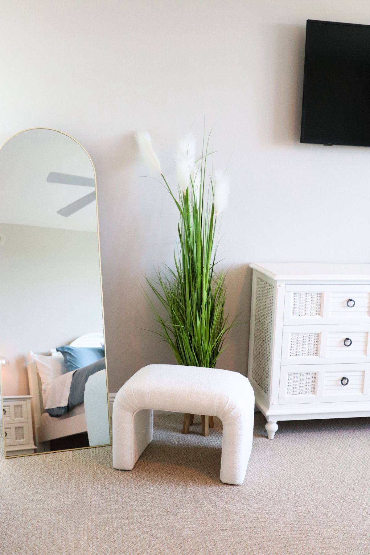 White ottoman, tall mirror, dresser, and green plant in a bright bedroom.