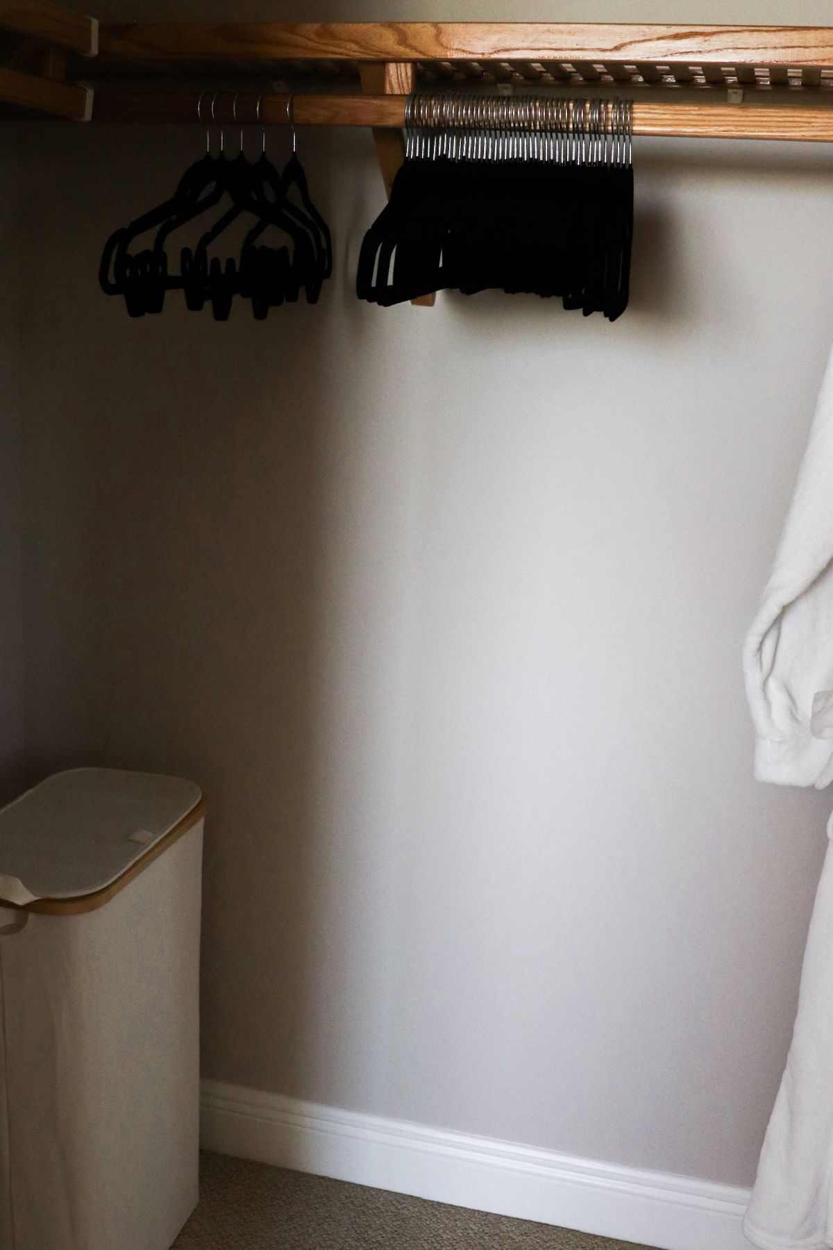 Closet with wooden shelving holding black hangers, laundry hamper, and robe.