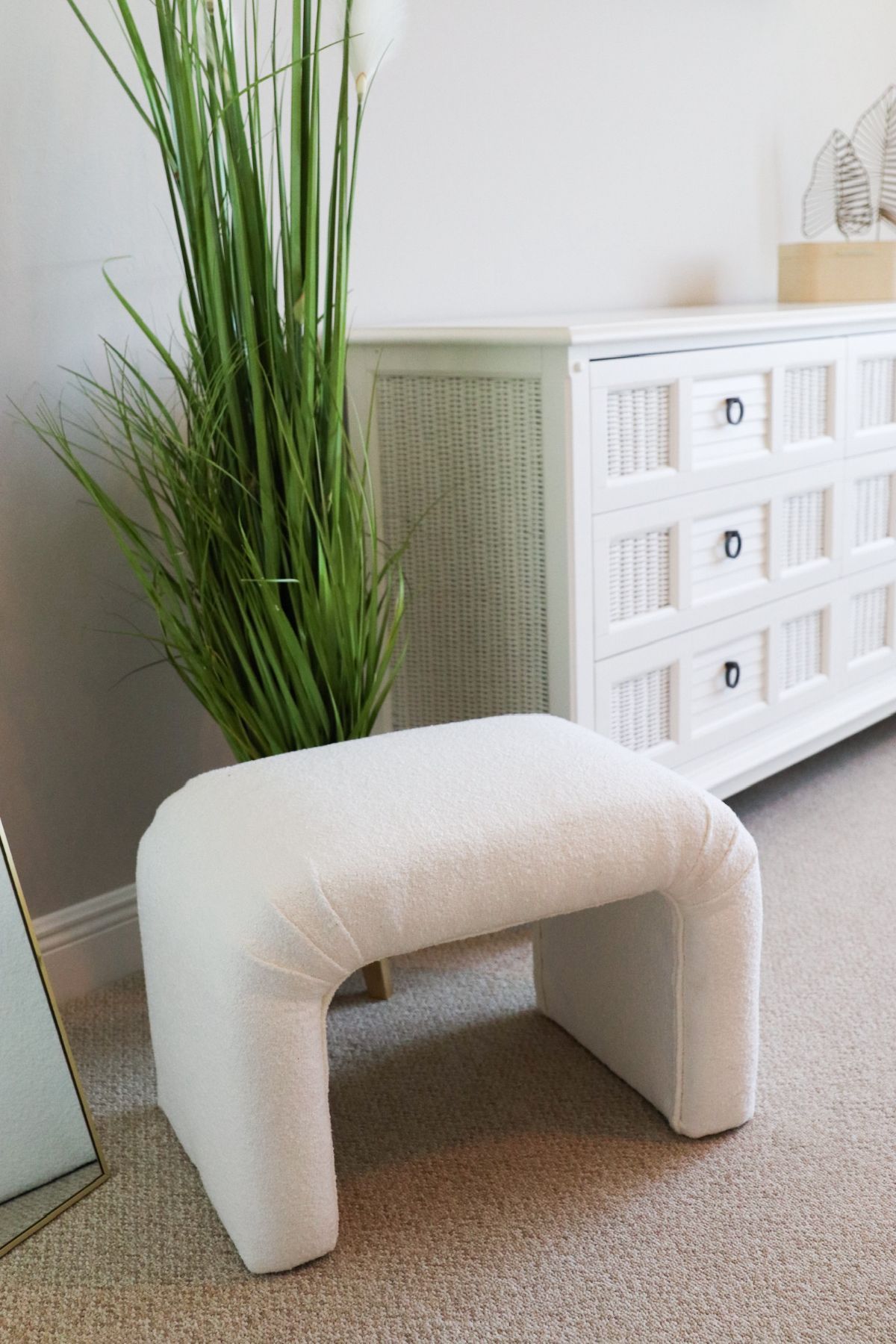 White upholstered ottoman in front of a white dresser and tall green plant on a carpeted floor.