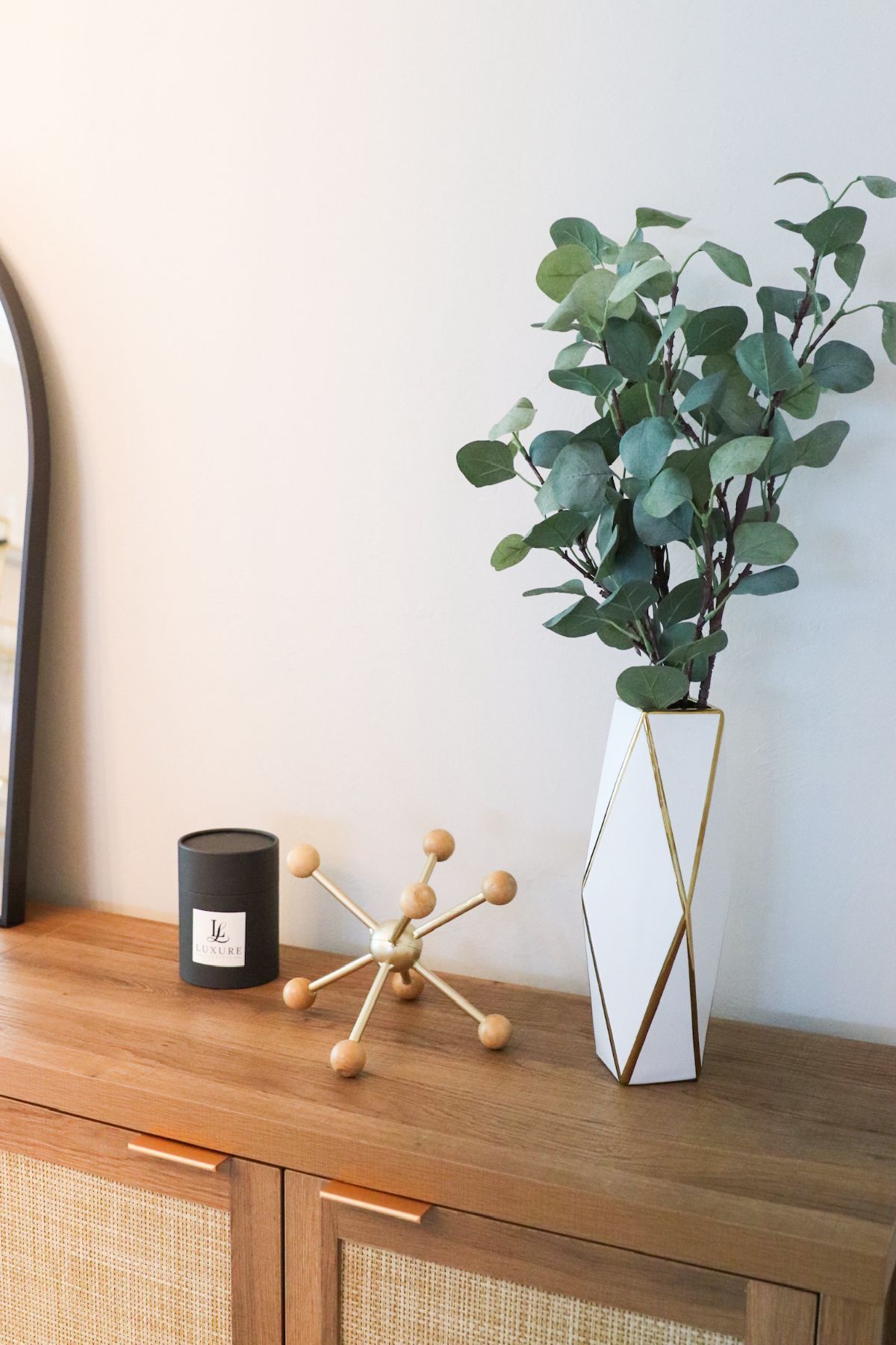 Wooden console table with decor: vase of eucalyptus, gold starburst, candle, and arched mirror.