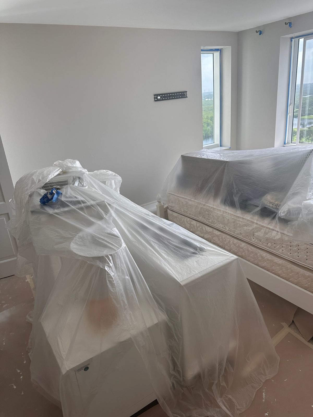 Room with white walls and furniture covered in plastic sheeting. Two windows offer an outside view.