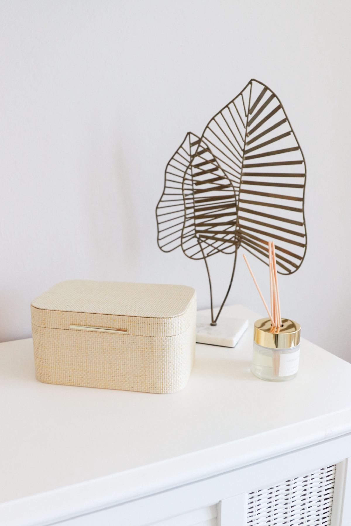 White dresser with woven box, decorative leaf sculpture, and reed diffuser.