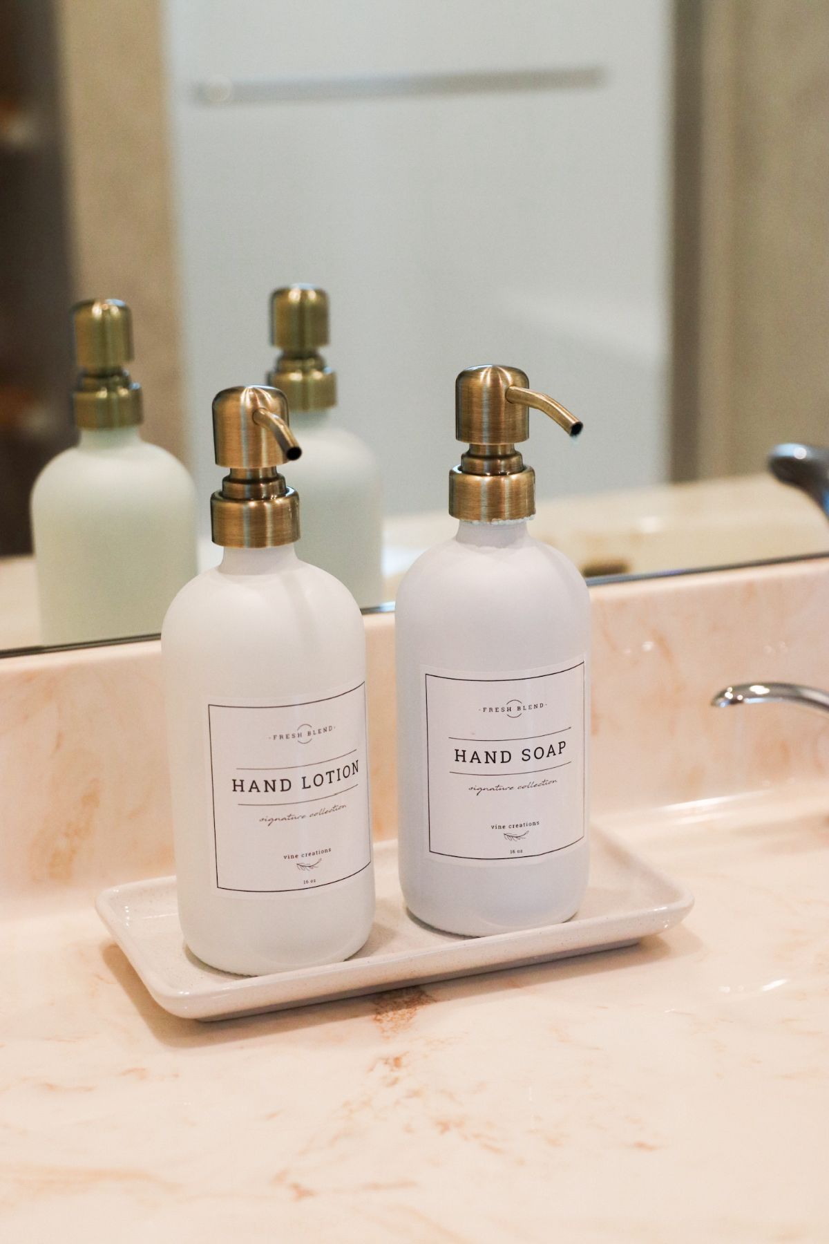 Two white soap dispensers with gold pumps on a tray, reflected in a mirror.