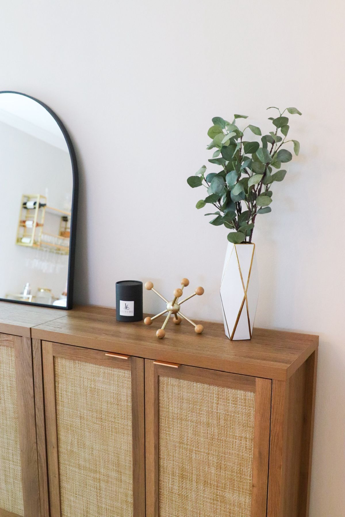 Wooden cabinet with vase of green leaves, black candle, and starburst decor. Black arched mirror in the background.