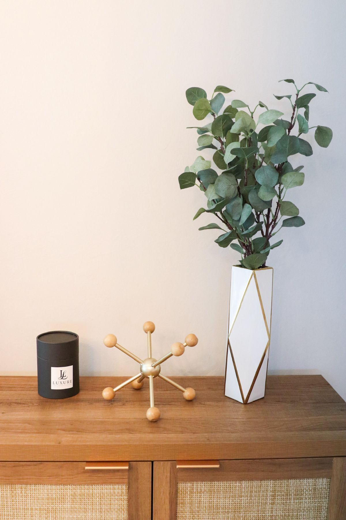 Wooden cabinet with decor: candle, starburst object, vase with eucalyptus against a beige wall.