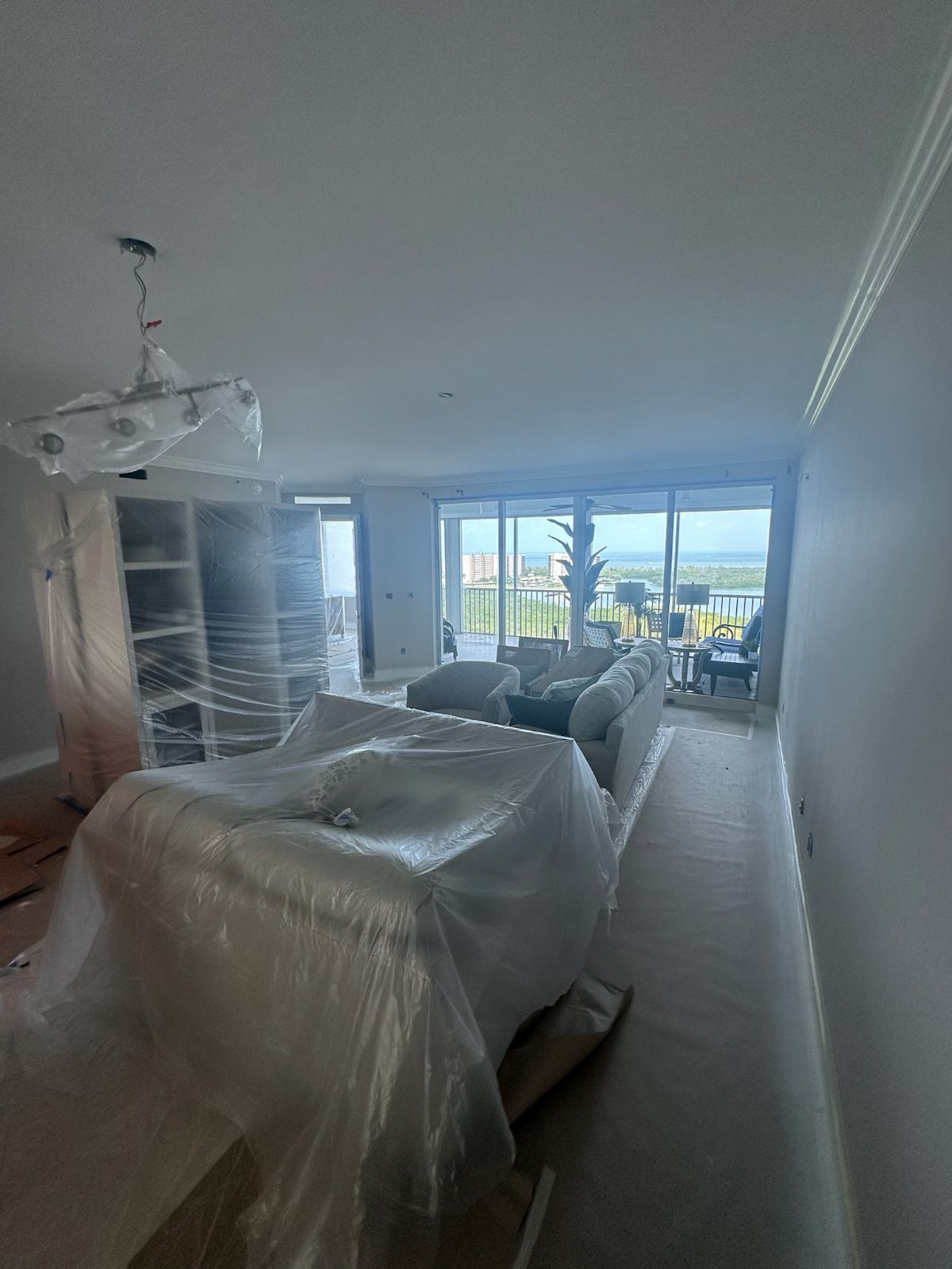 Room with furniture wrapped in plastic, with large windows overlooking a scenic view.