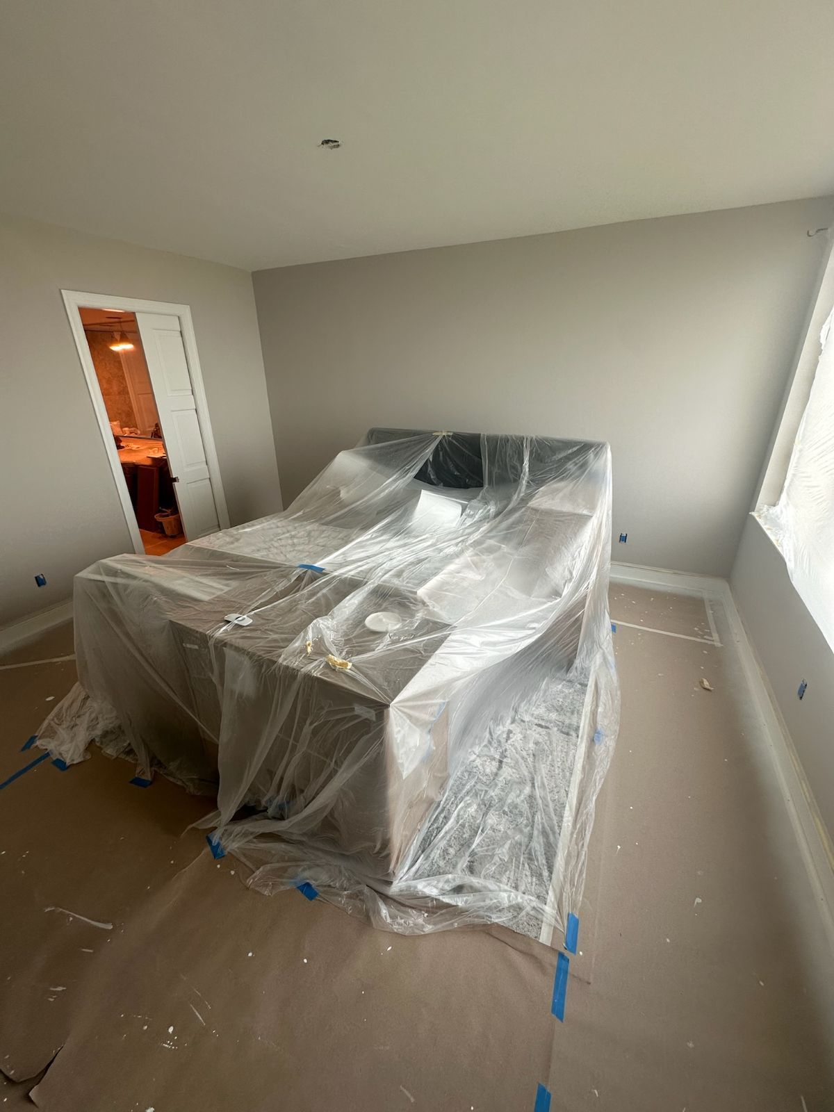 Room with a bed covered in plastic, likely during construction or renovation. Pale walls, wood floors.