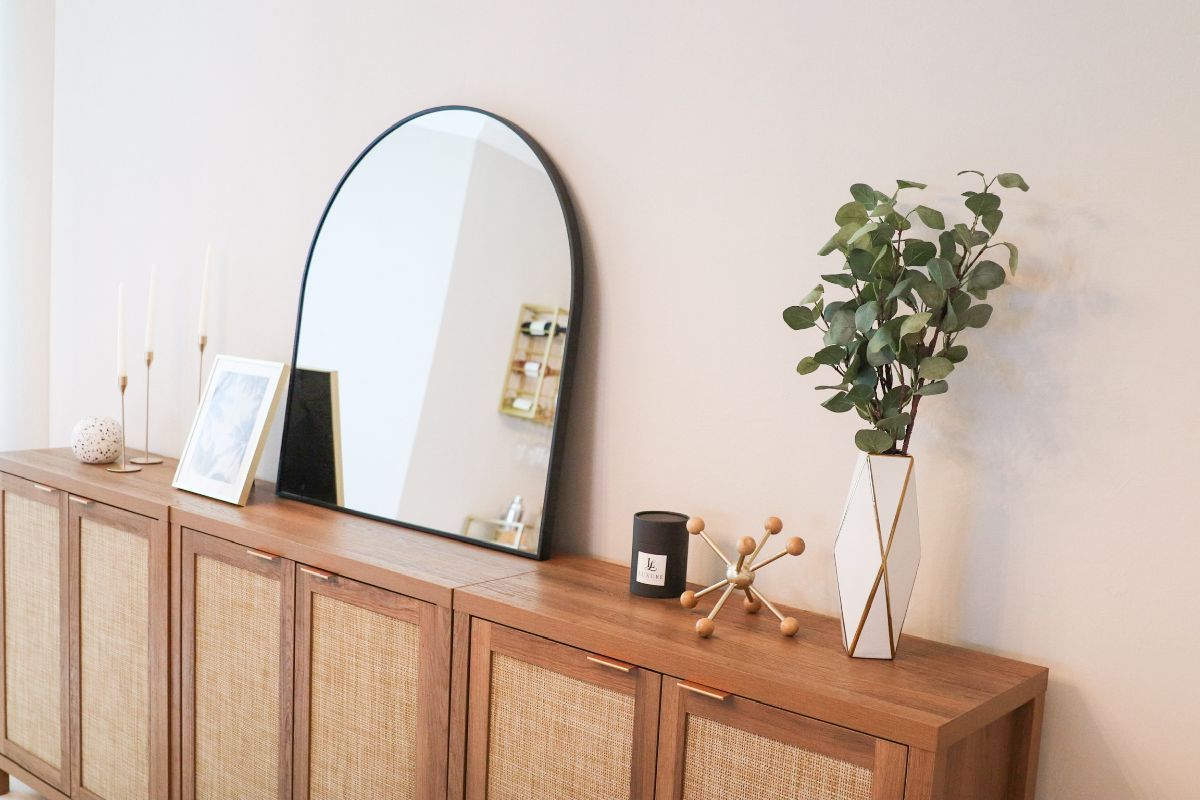 Wooden sideboard with arch mirror, greenery, and decor against a light pink wall.