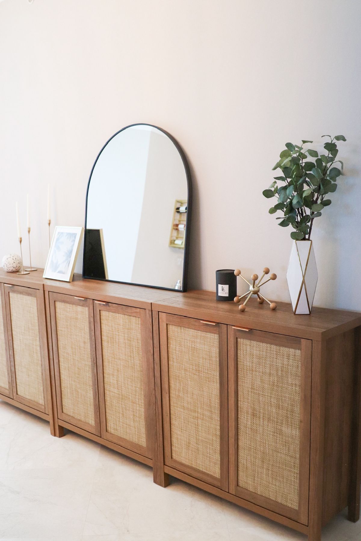 Wooden cabinet with woven panels, arched mirror, vase of greenery, and decorative items.