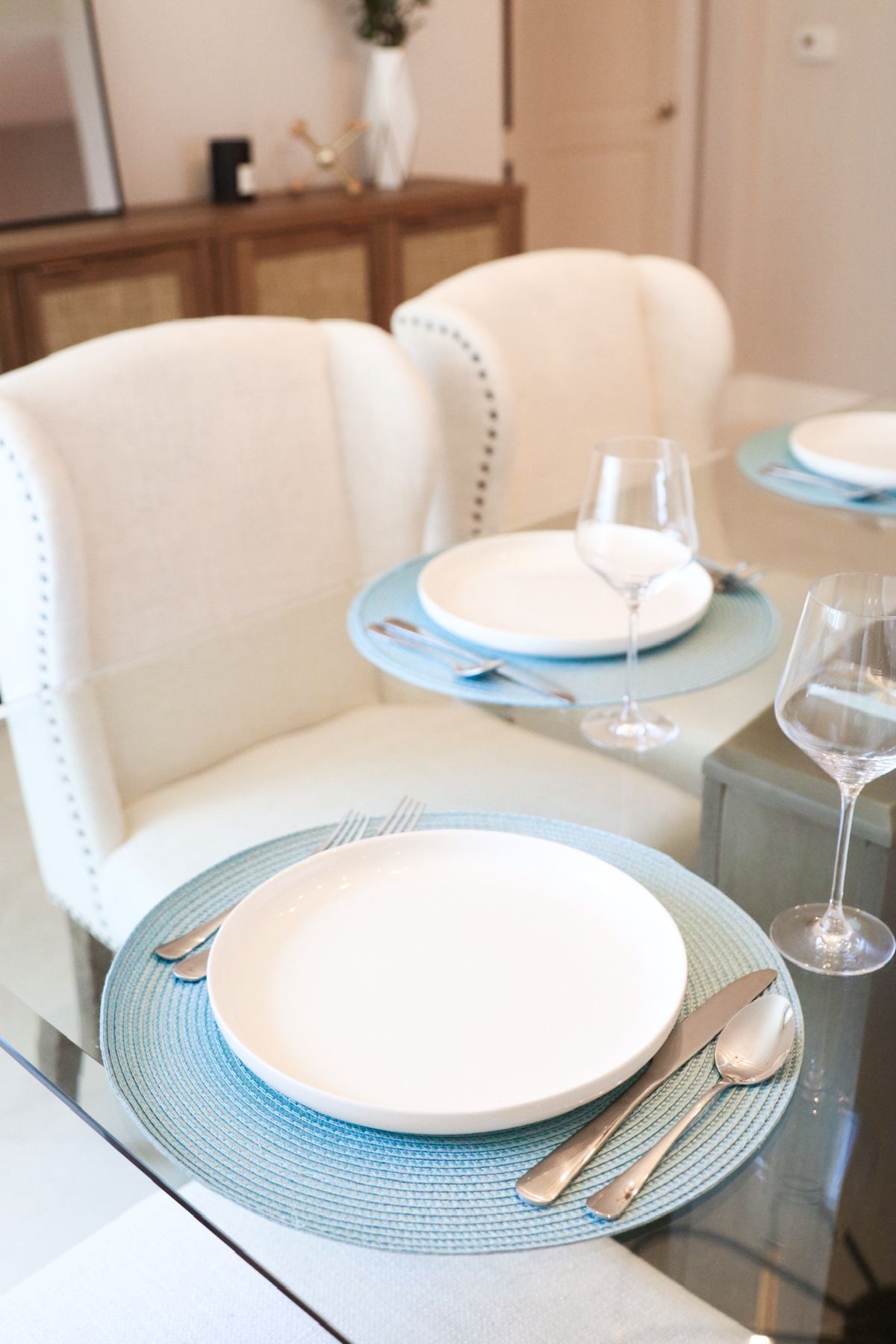 Dining table set with white plates, silverware, and wine glasses. Blue placemats and white chairs.