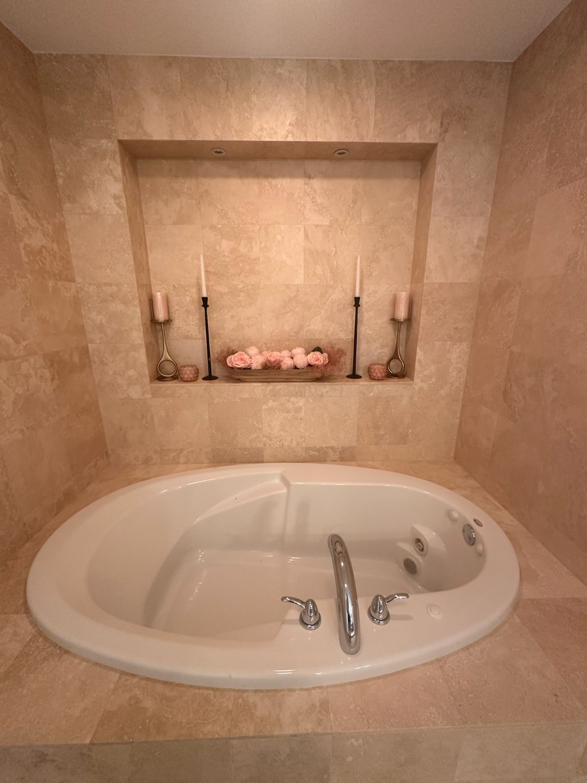 Oval bathtub, with a recessed shelf above holding candles and flowers, surrounded by beige tile.