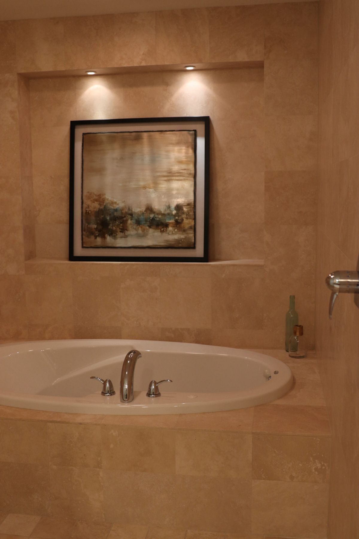 Oval bathtub built into a beige tiled wall with framed artwork and recessed lighting above.