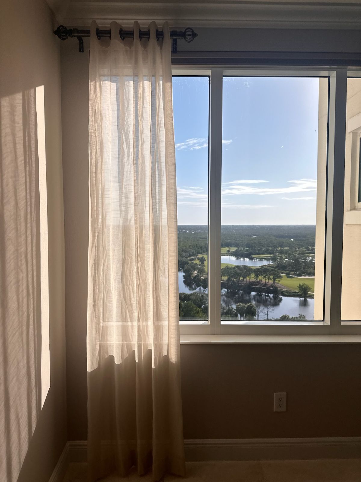 A sheer beige curtain hangs next to a window overlooking a landscape with water and trees under a blue sky.