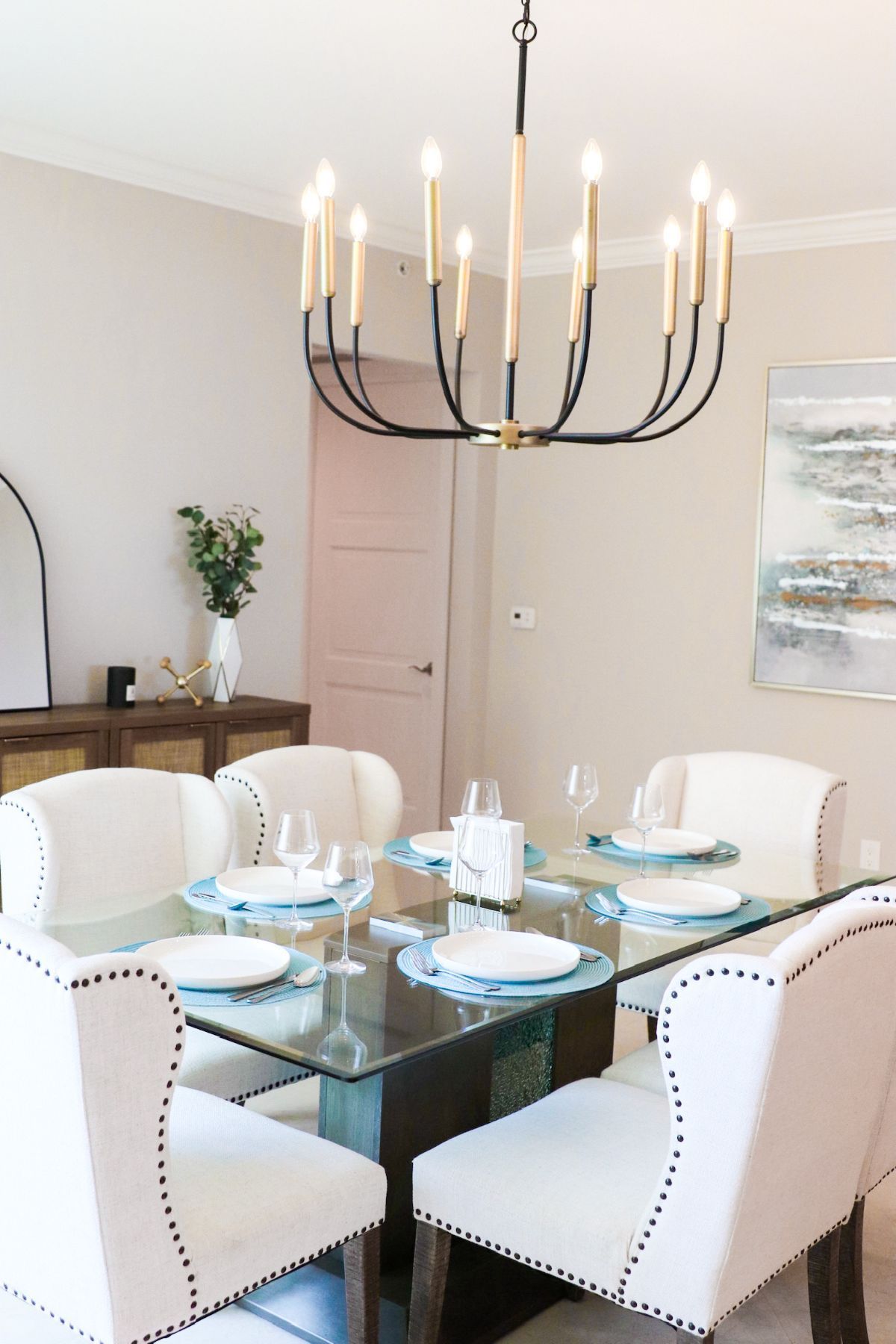 Dining room with a glass table set for a meal, chandelier overhead, white chairs, and artwork on the wall.