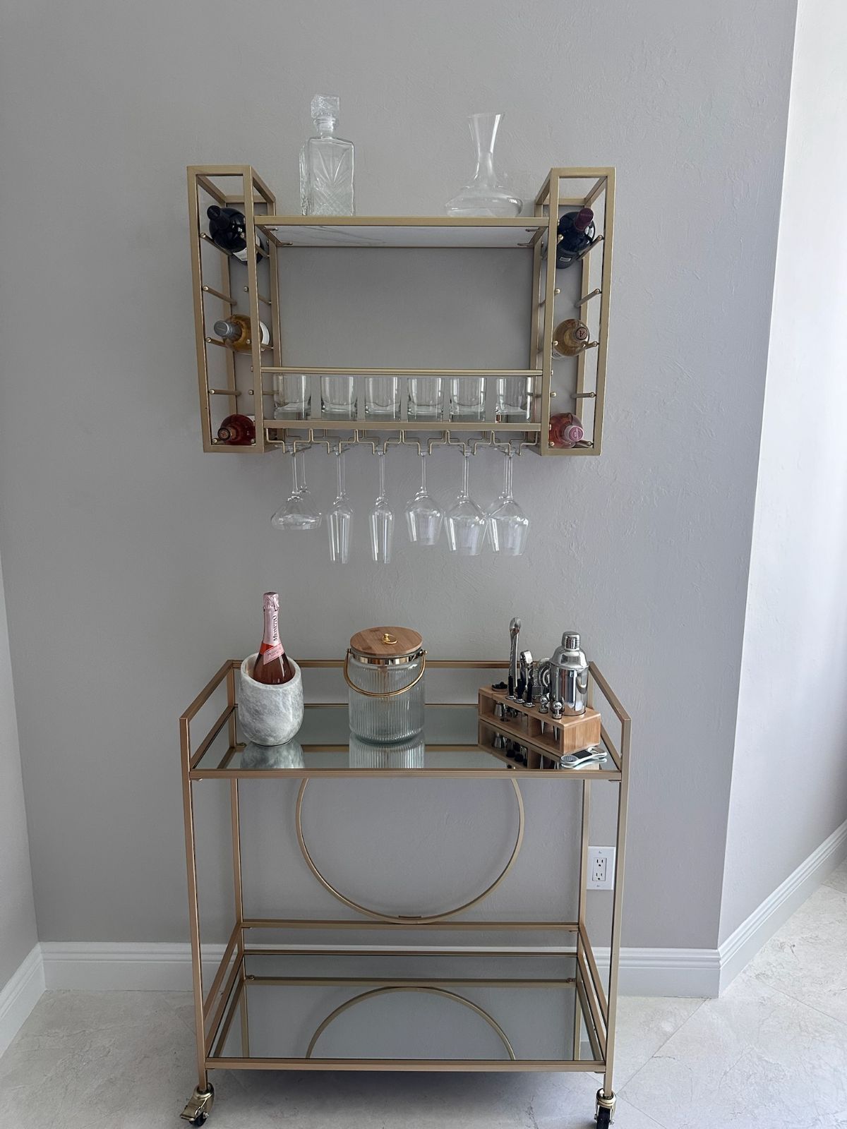 Gold bar cart and wall-mounted wine rack against a gray wall. Contains bottles, glasses, and barware.
