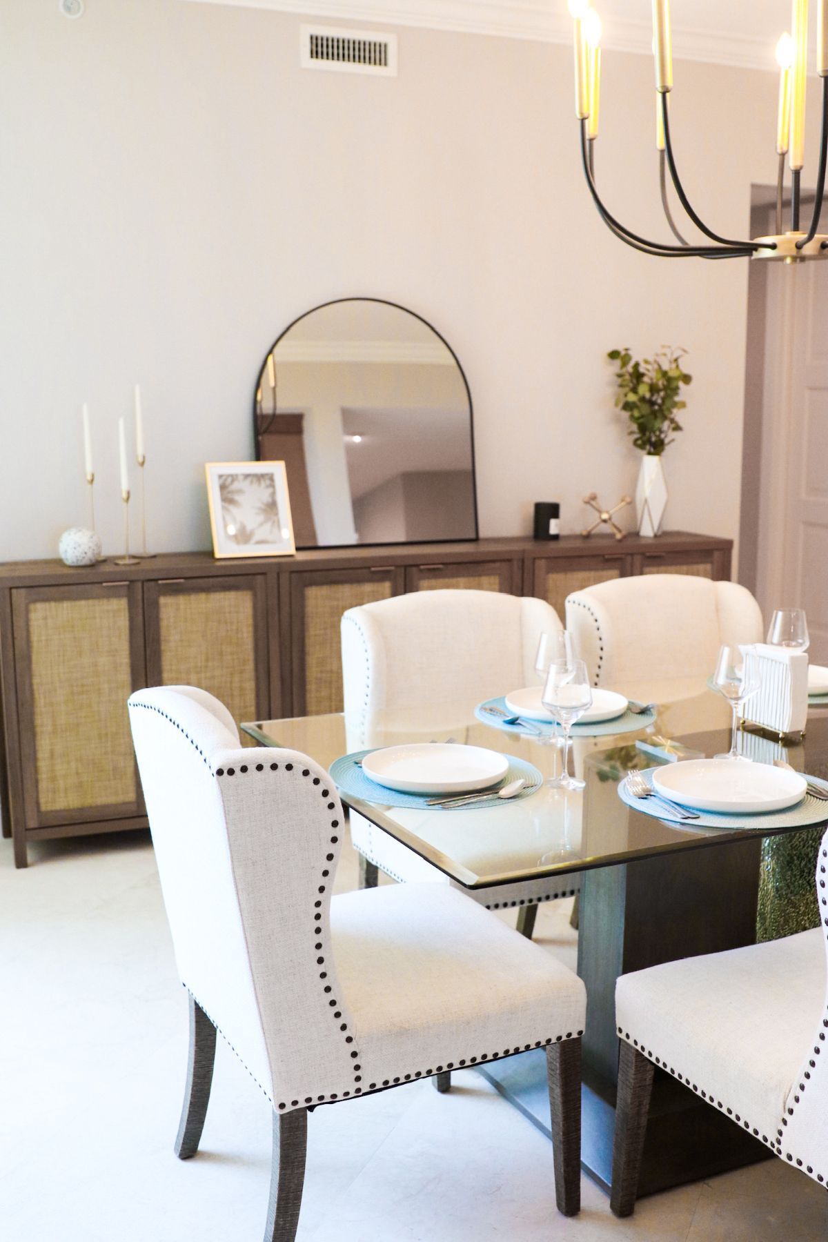 Dining room with wood buffet, round mirror, glass table, white chairs, and candle chandelier.
