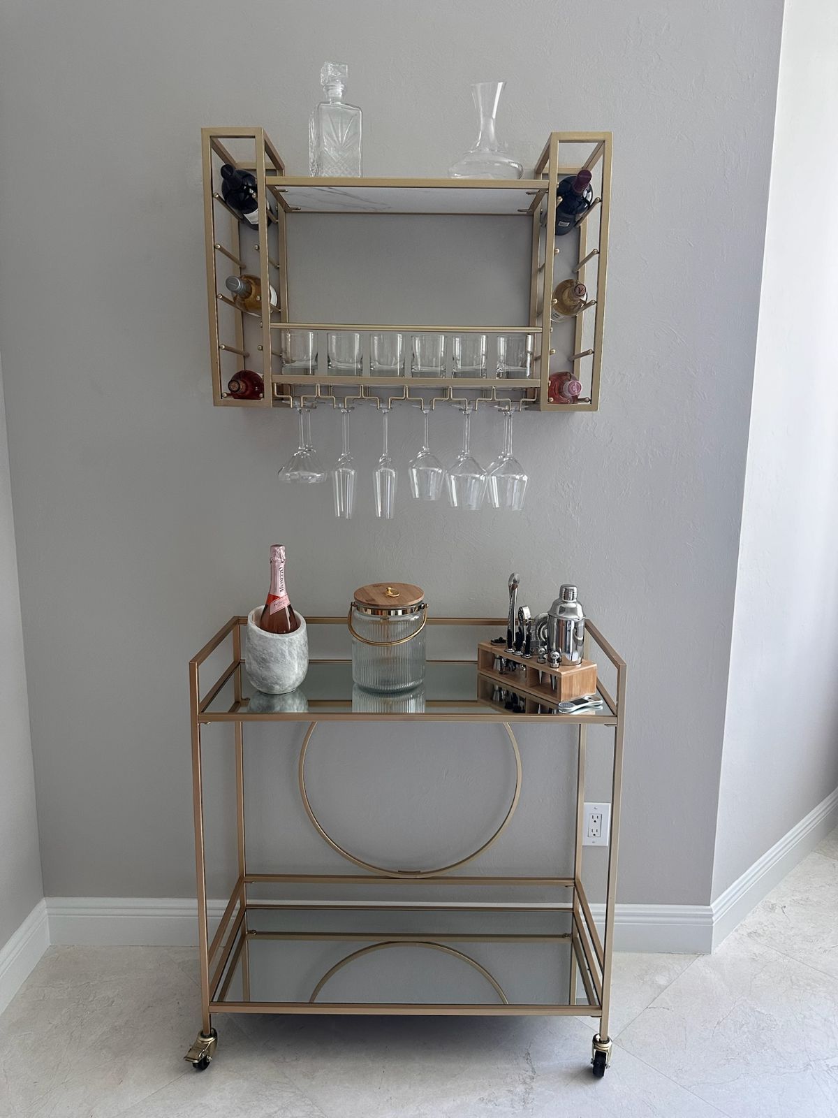 Gold bar cart and wall-mounted wine rack against a gray wall. Wine bottles, glasses, and barware are displayed.