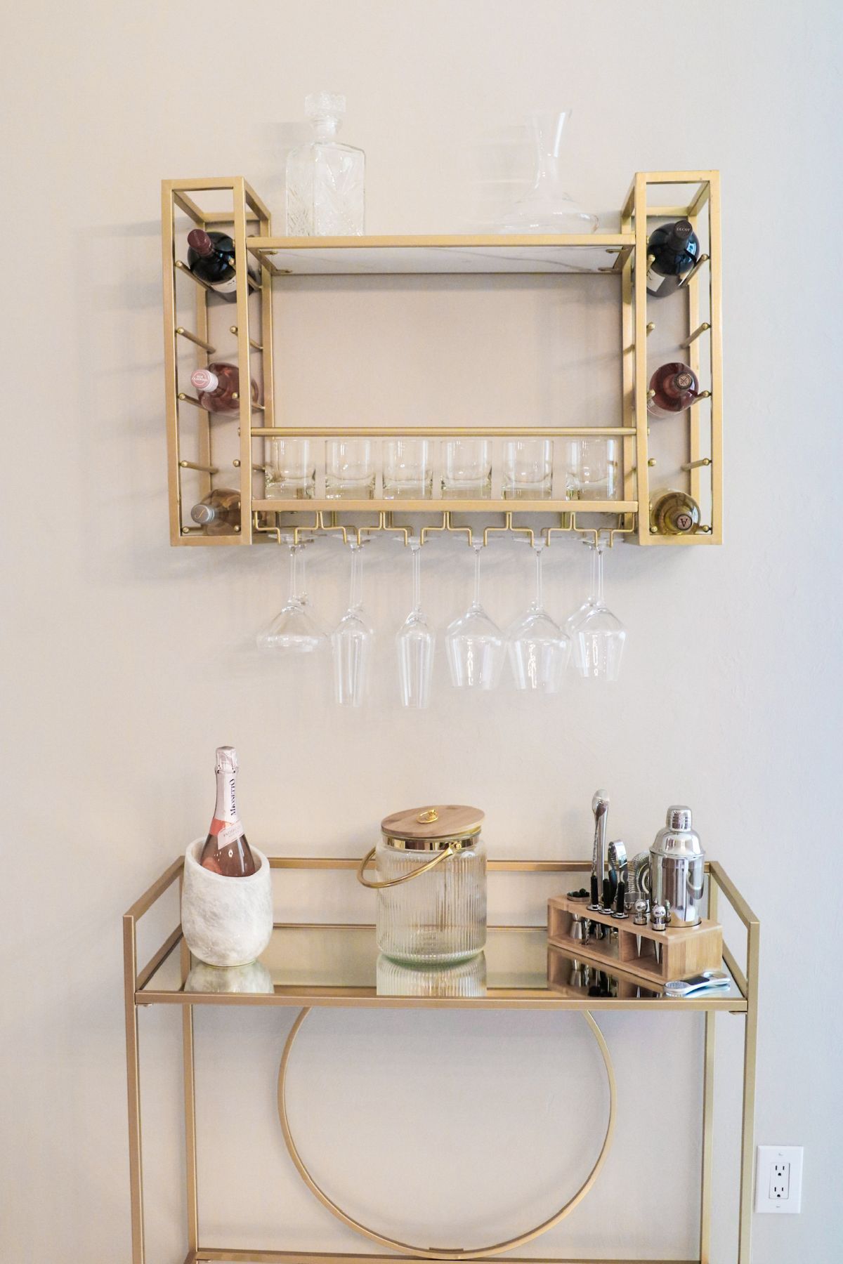 Gold bar cart and wall rack with glassware and bottles against a neutral wall.