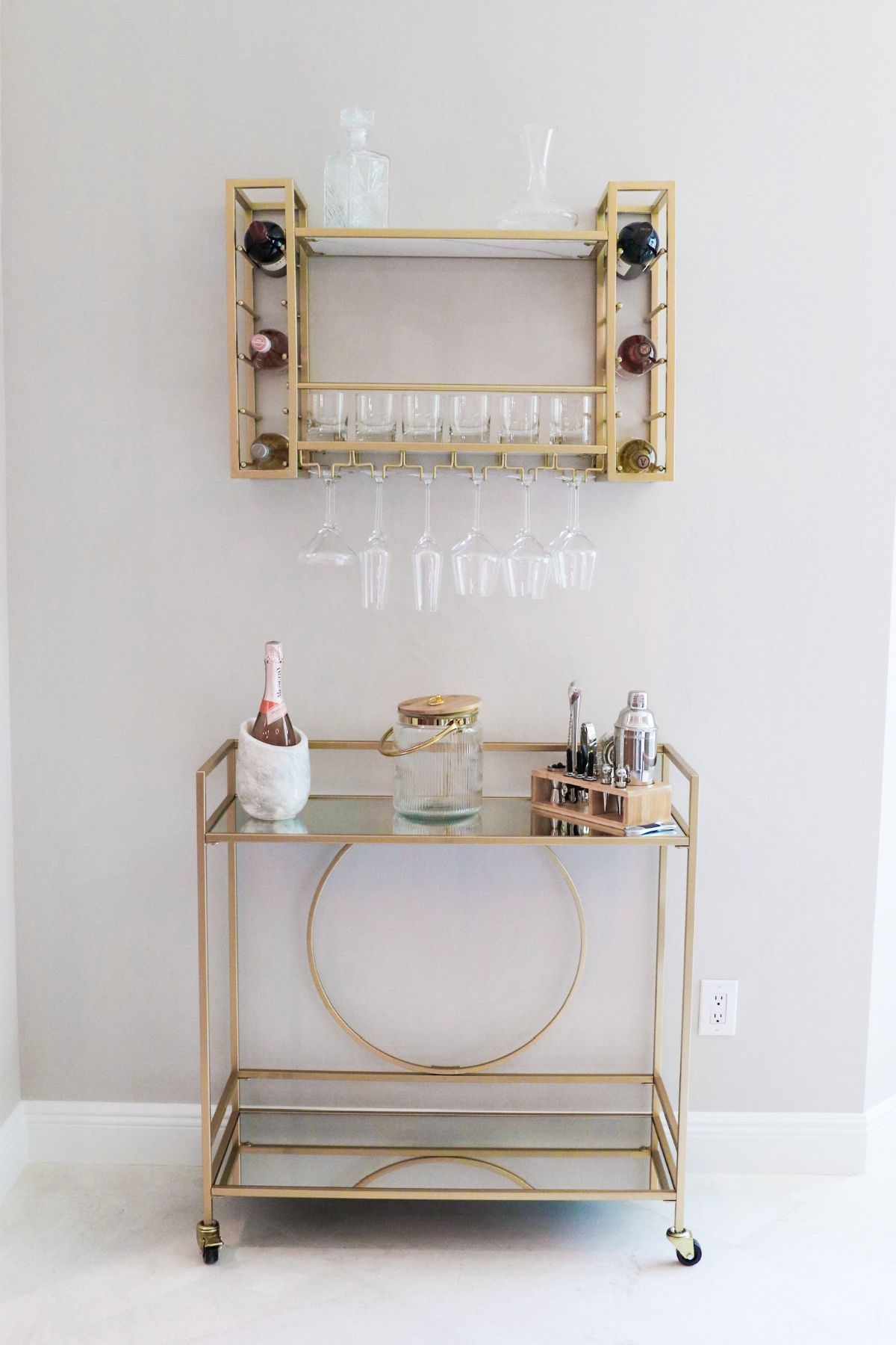 Gold bar cart and wine rack on a light wall. Champagne bottle, glasses, and barware.