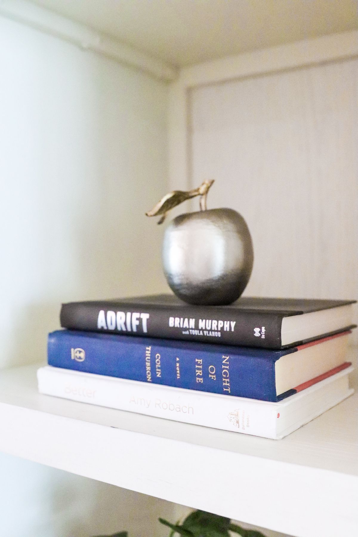 Books stacked with silver apple ornament on a white shelf.