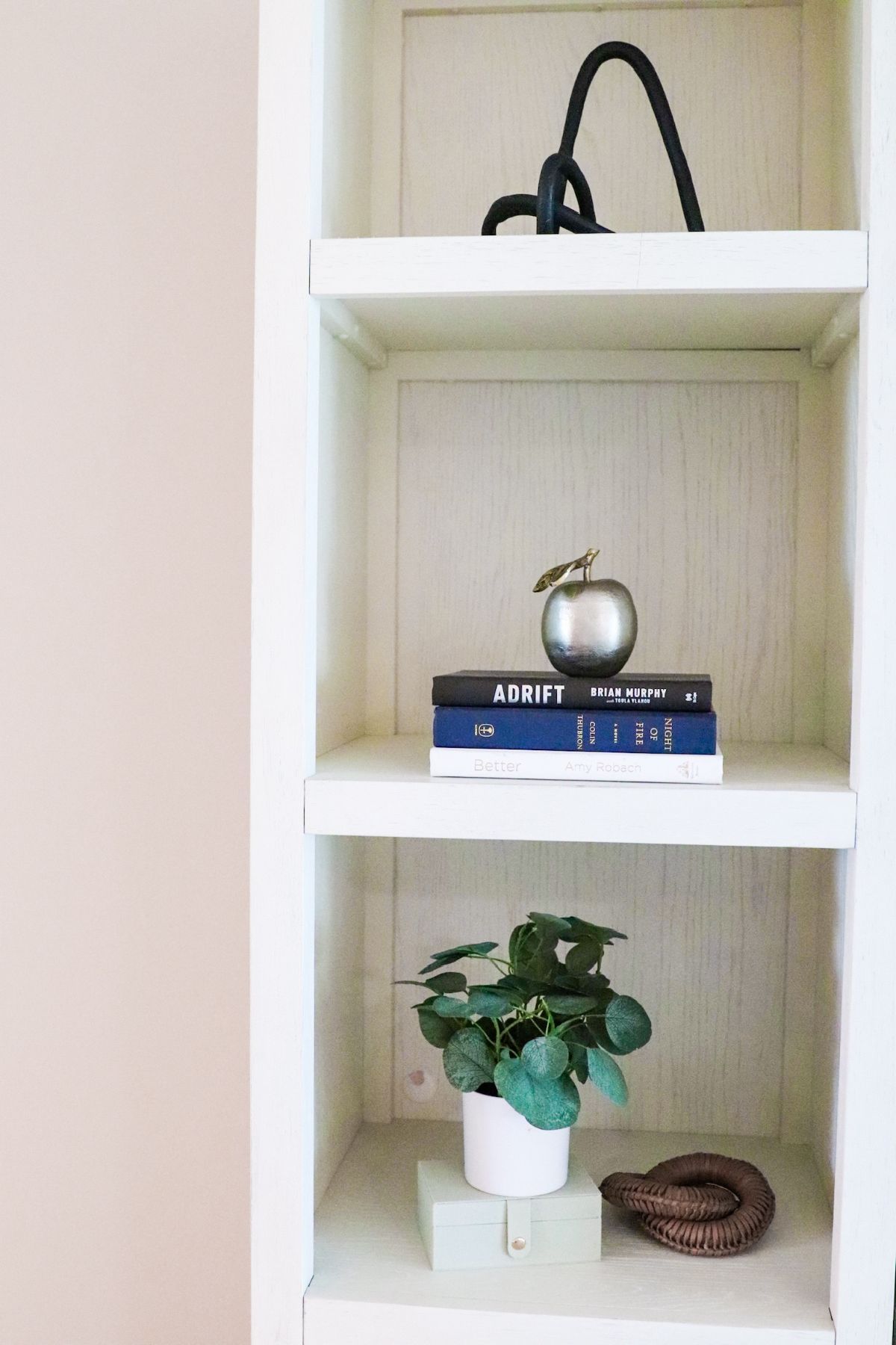 White bookshelf with decorative items: a black sculpture, silver apple, books, plant, and wooden coil.