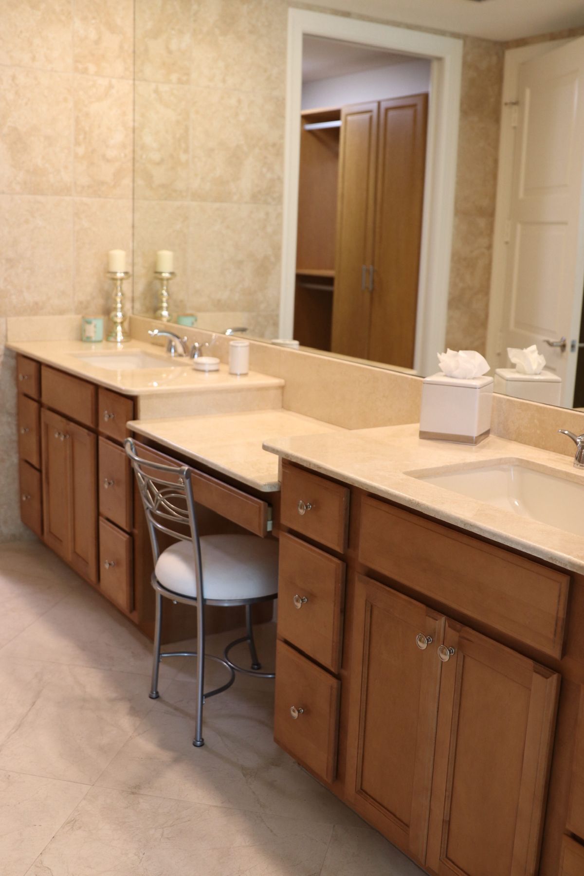 Bathroom with light-colored cabinets, countertops, and a silver chair. A large mirror and a walk-in closet are visible.