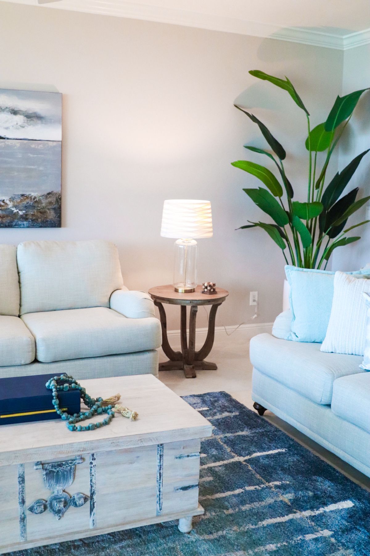 Living room with cream sofas, wooden side table with lamp, large plant, blue rug, and trunk-like coffee table.