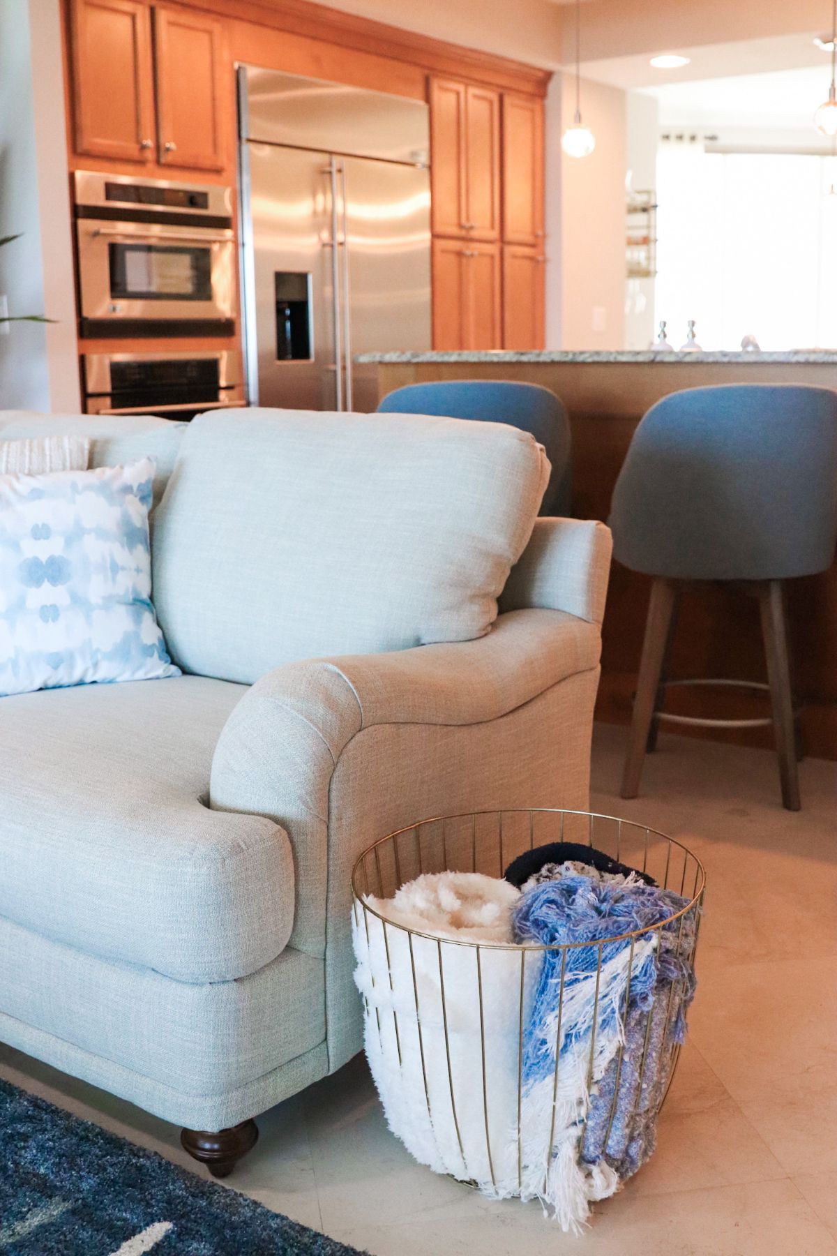 Living room with a light-colored couch, a metal basket holding folded textiles, and a stainless steel refrigerator.