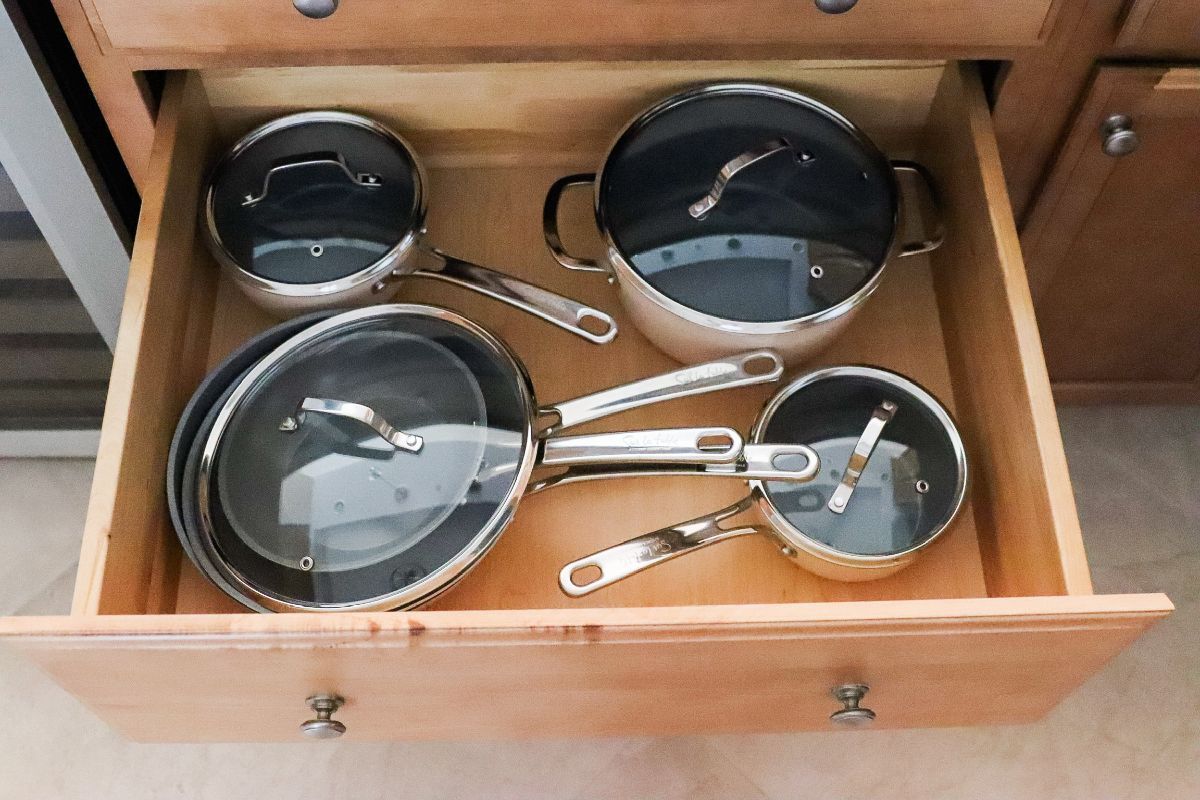 A drawer holding five pots and pans with glass lids, inside a wooden cabinet.