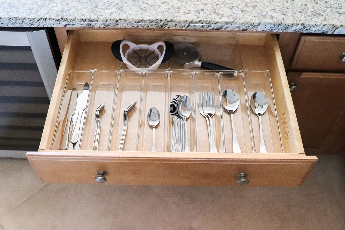 A kitchen drawer with organized silverware in transparent dividers, plus a heart-shaped object and a pizza cutter.