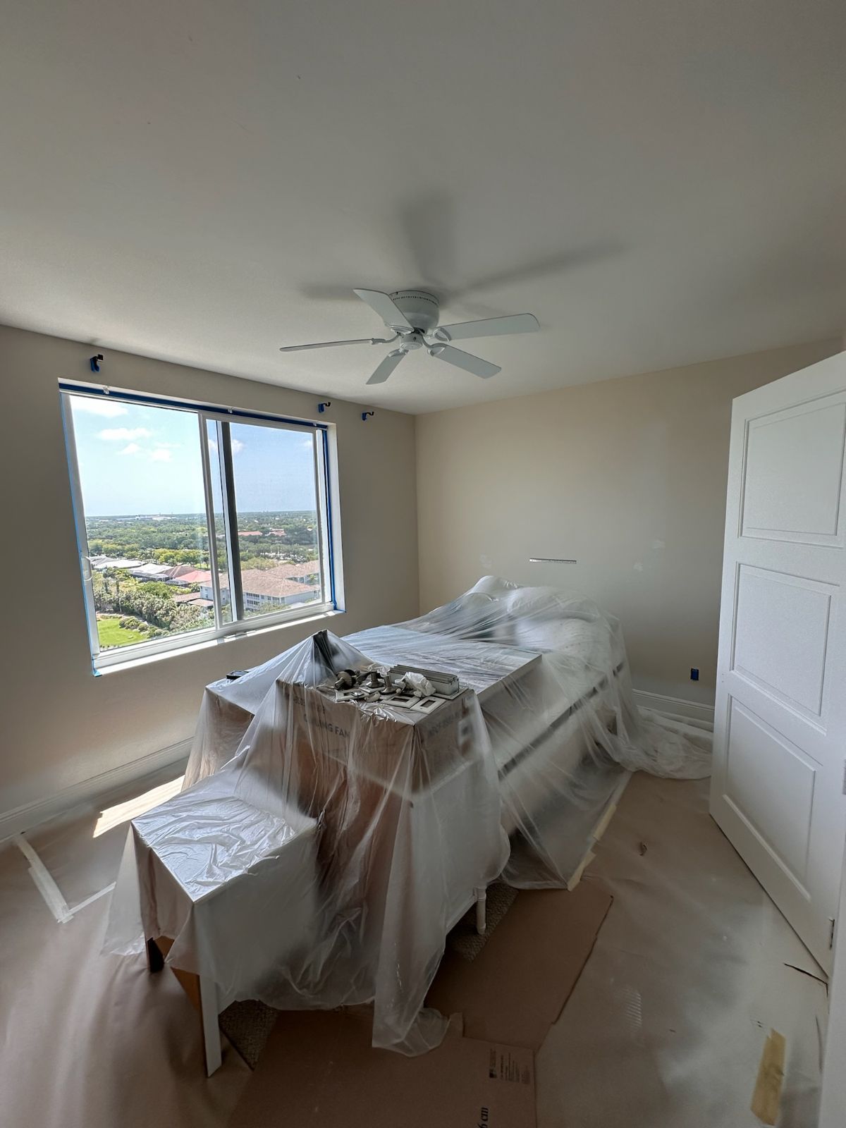 Bedroom with furniture covered in plastic for renovation. Light walls, window, ceiling fan, and white door.