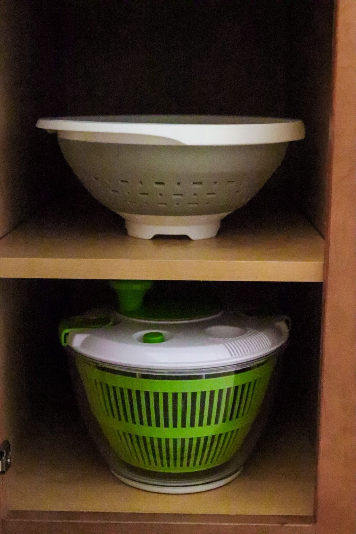 A green salad spinner and a white colander sit on shelves inside a cabinet.