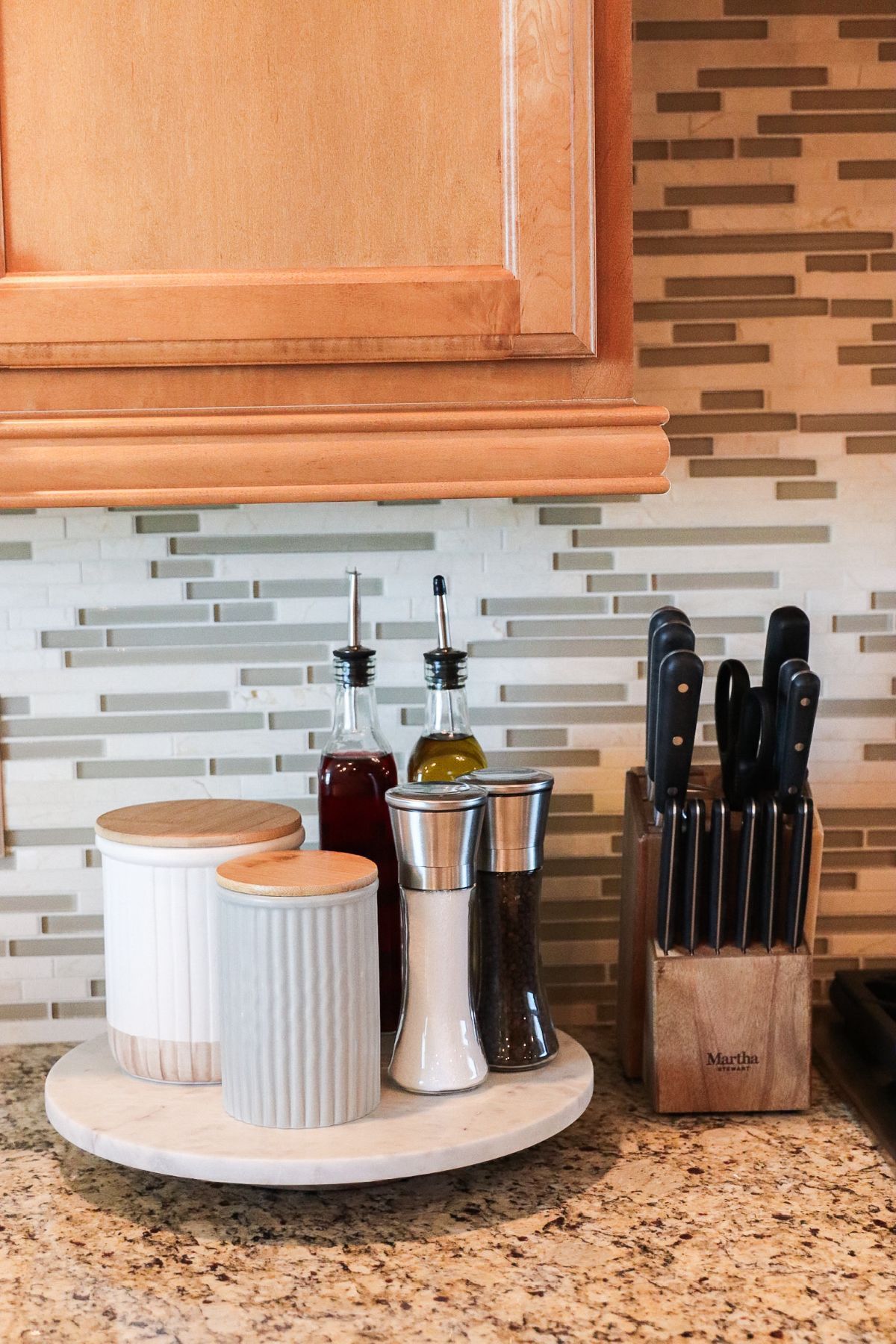 Kitchen countertop with jars, oil bottles, salt & pepper shakers, and a knife block on a lazy susan, near a backsplash.