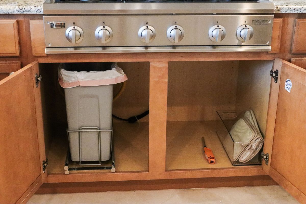 Cabinet under a stove with a trash can and baking pan. The cabinet doors are open.