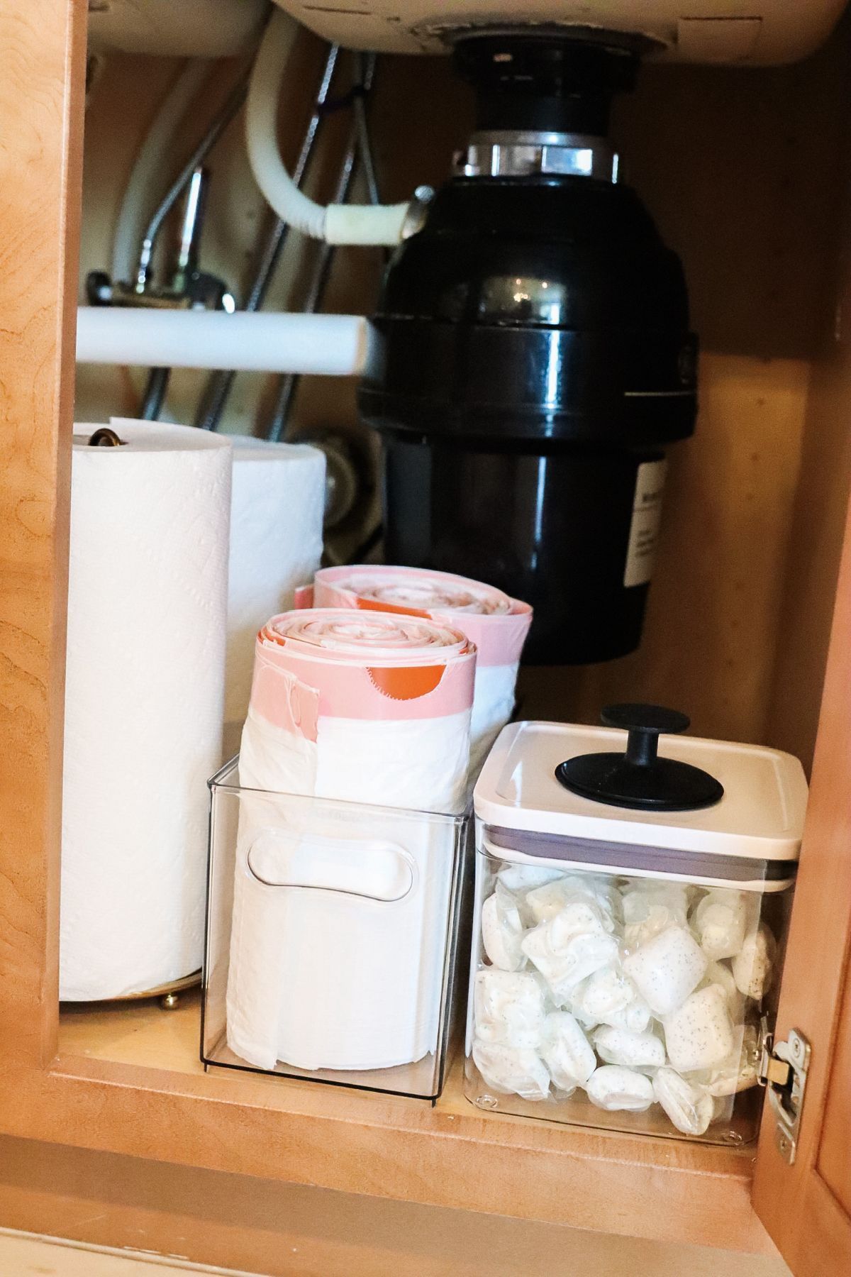 Under-sink cabinet with garbage disposal, paper towels, and containers holding items.