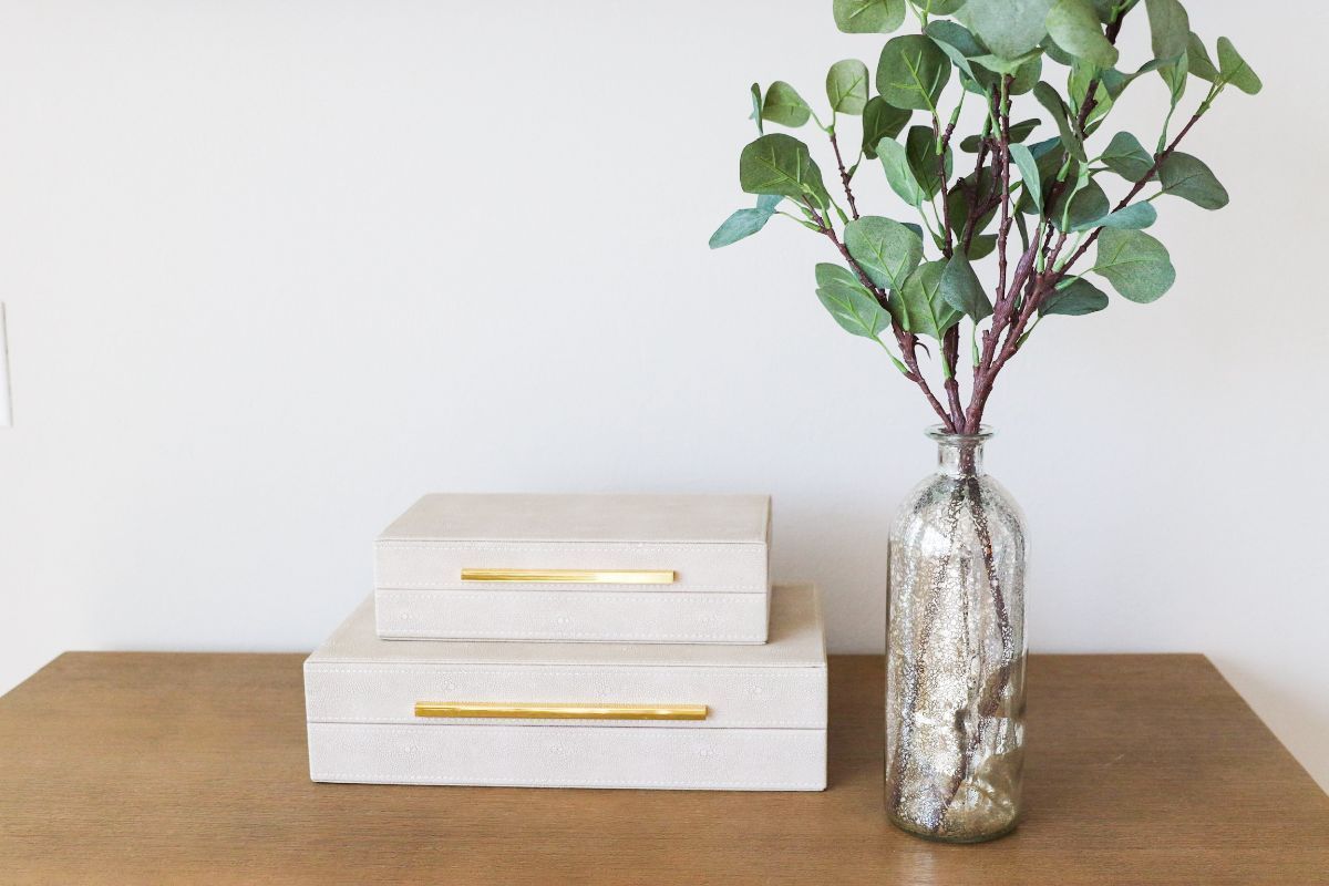 Two beige boxes with gold handles and a vase of greenery on a wooden surface.