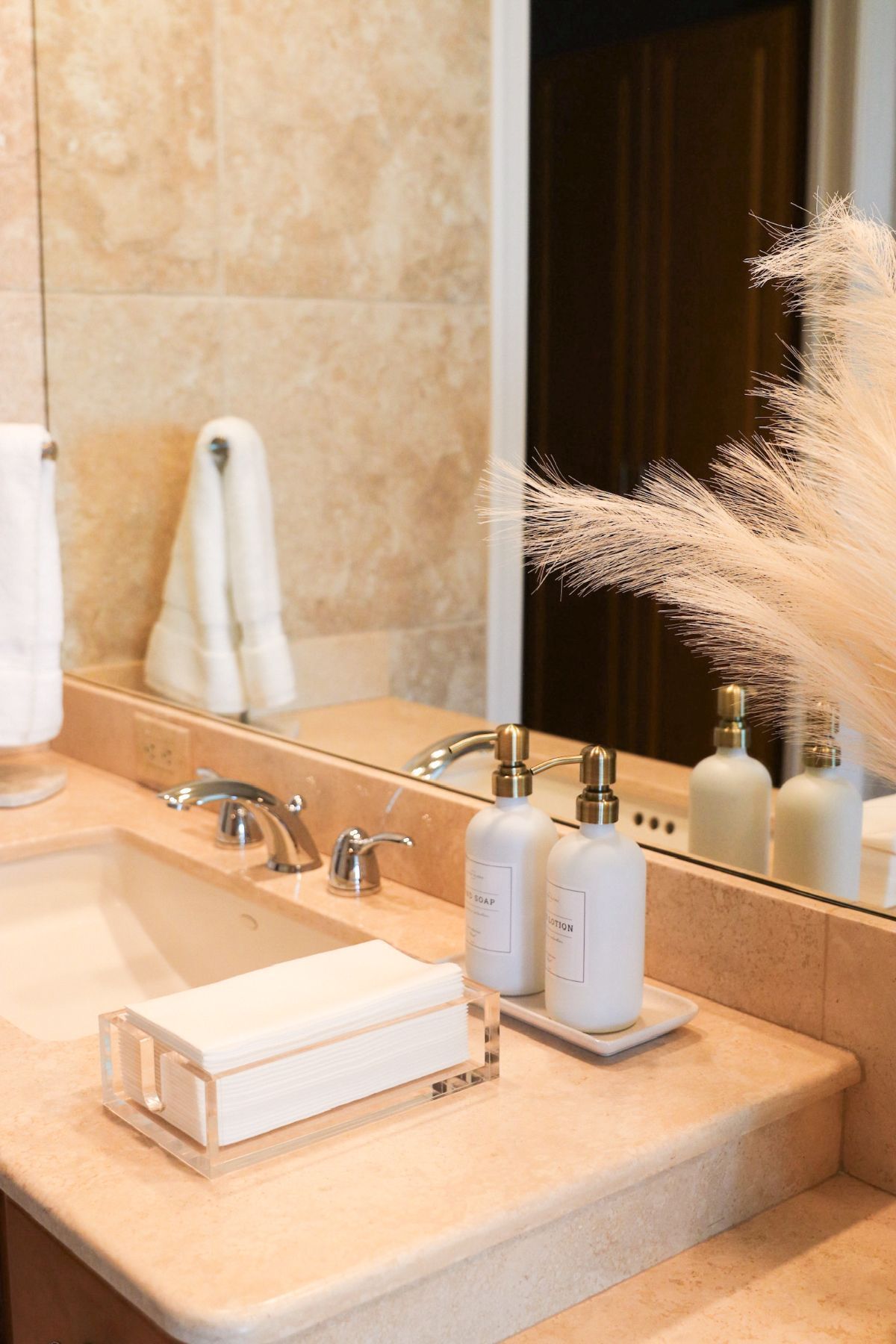Bathroom vanity with beige countertop, sink, mirror, and toiletries. Towels and pampas grass decorate the space.