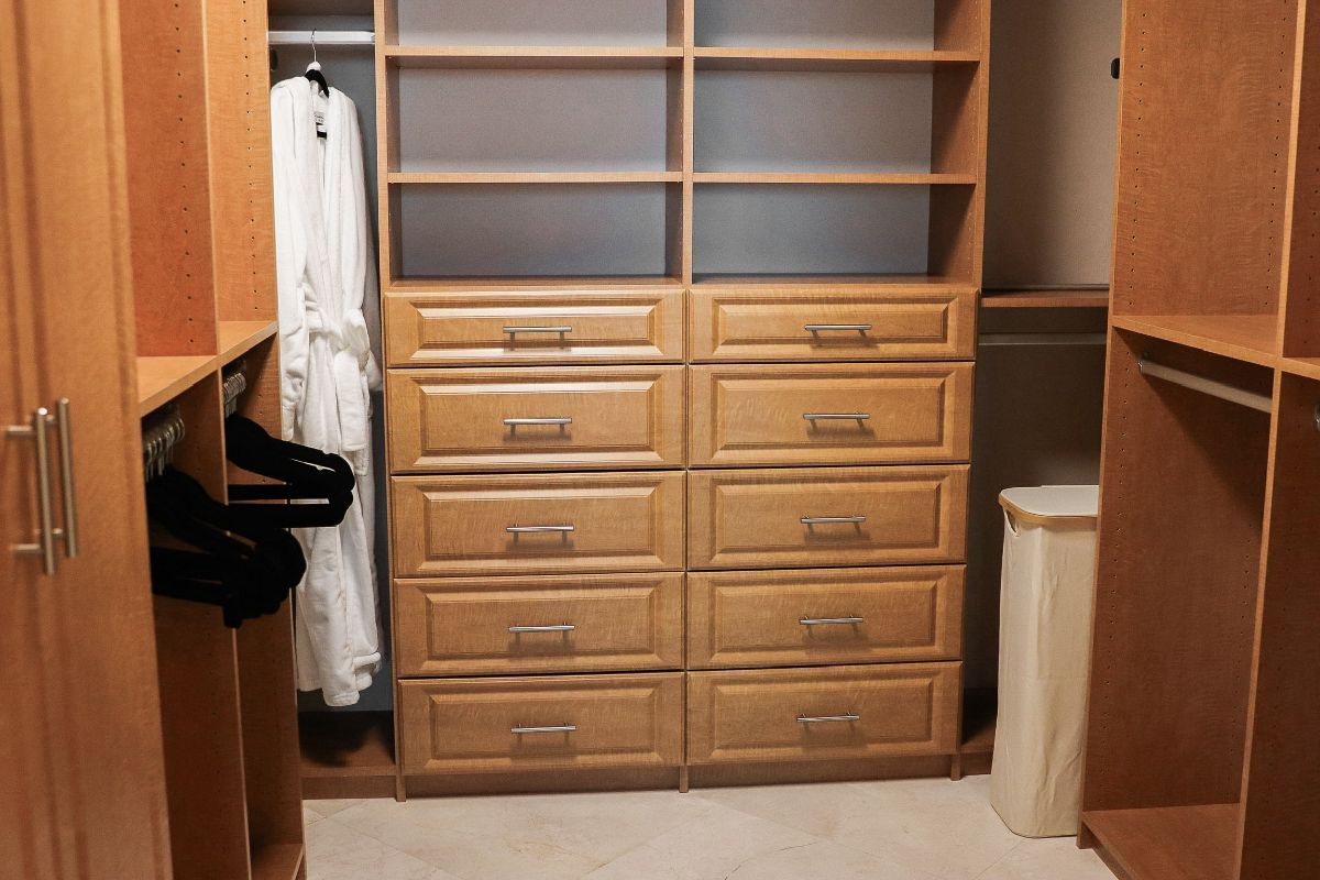Walk-in closet with light brown wood shelves, drawers, and hanging rods. A bathrobe hangs on a rod.