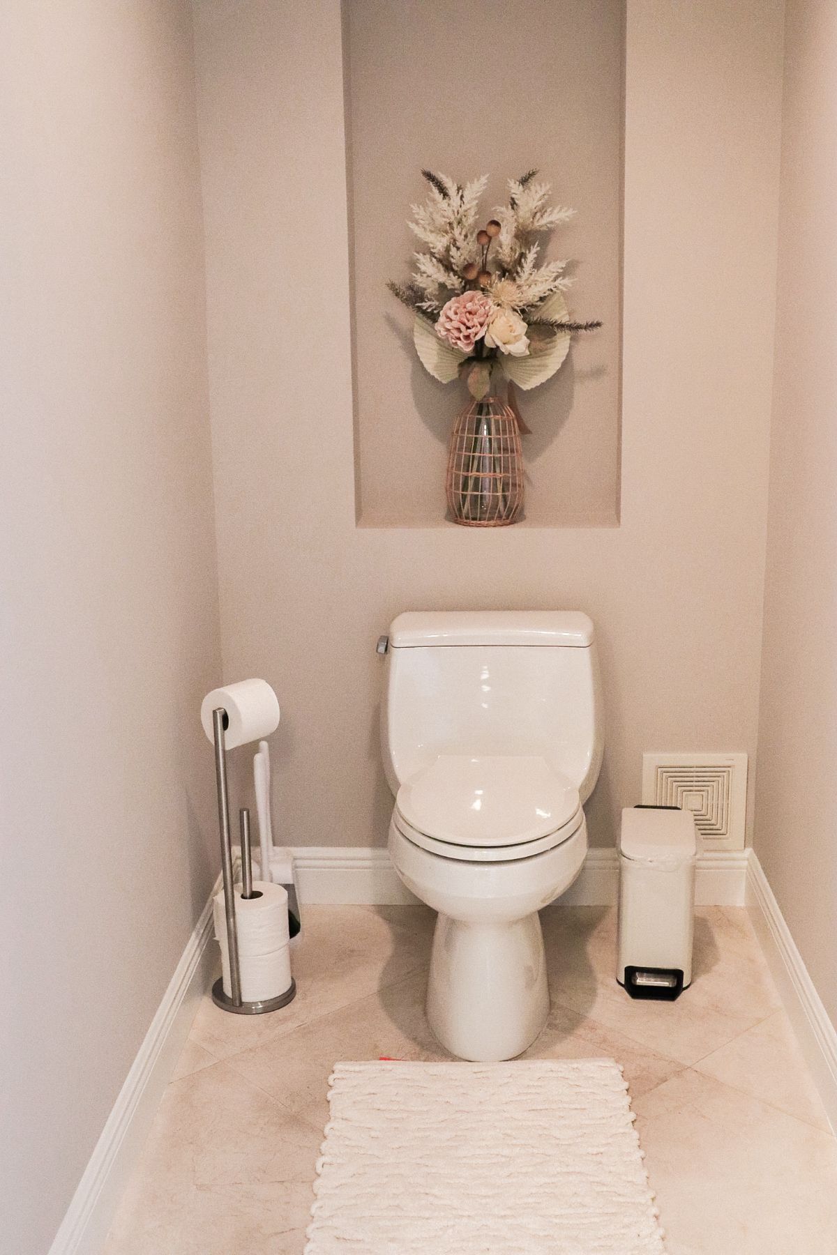 White toilet in a small bathroom with neutral walls, floral arrangement, and a rug.