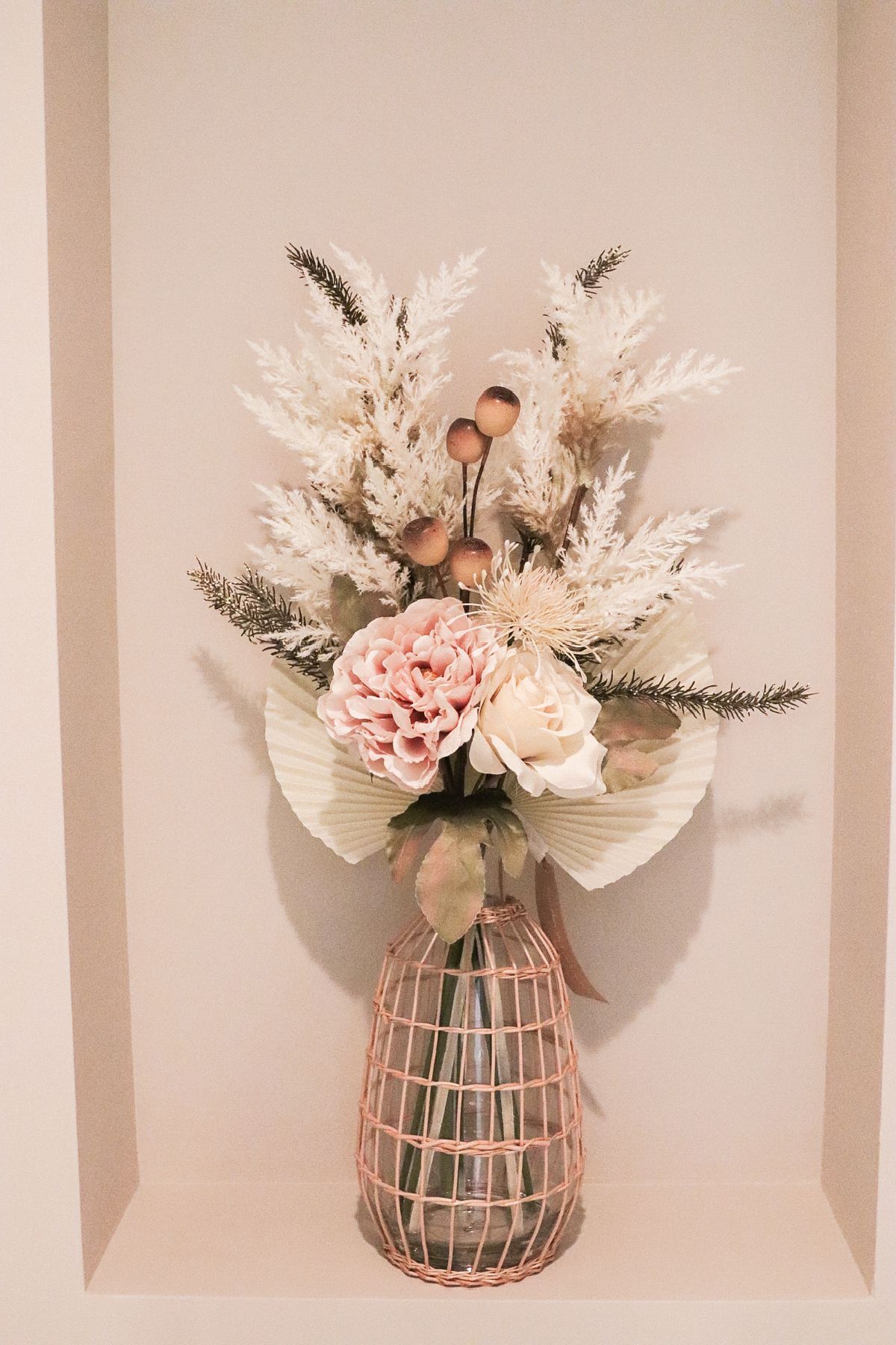 Pink and cream floral arrangement in a rose gold wire cage vase, set in a recessed wall niche.